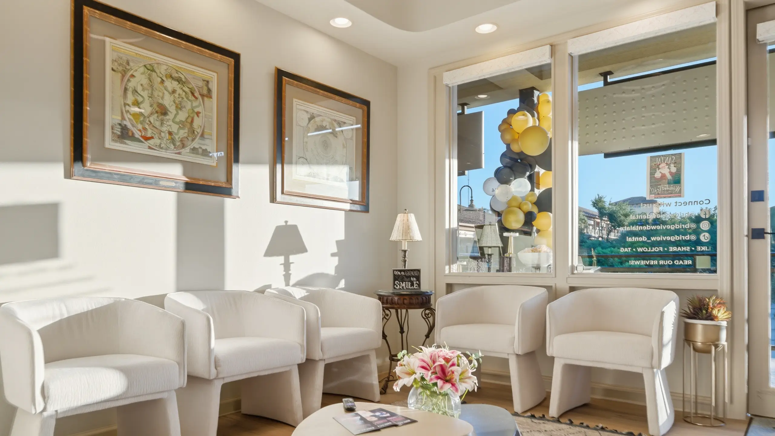Modern waiting room with five white armchairs, a round coffee table with flowers, framed maps on the wall, and large windows showing balloons outside.