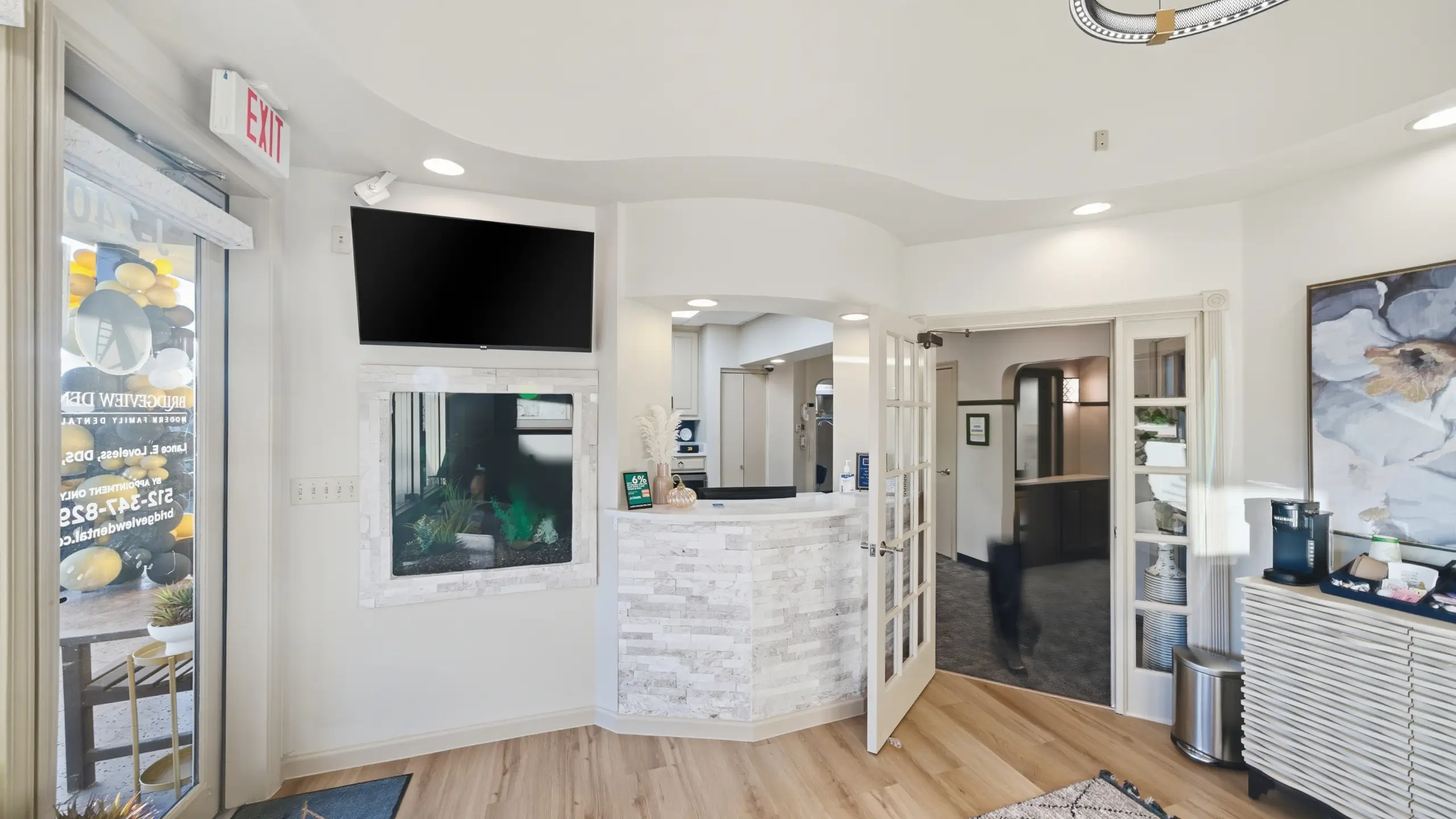 Modern dental office reception area with a white stone front desk, glass door entry, mounted TV, and a coffee station.