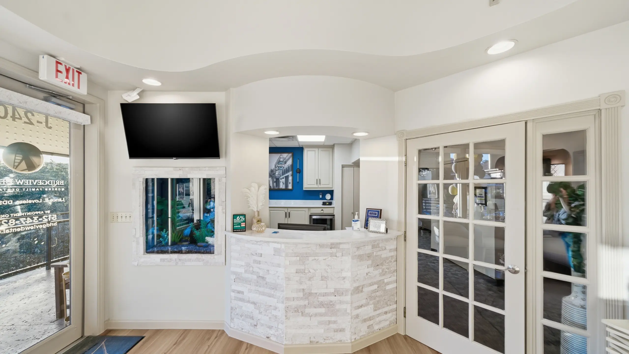 Reception area with white stone front desk, mounted TV above a small aquarium, and glass panel double doors to the right.