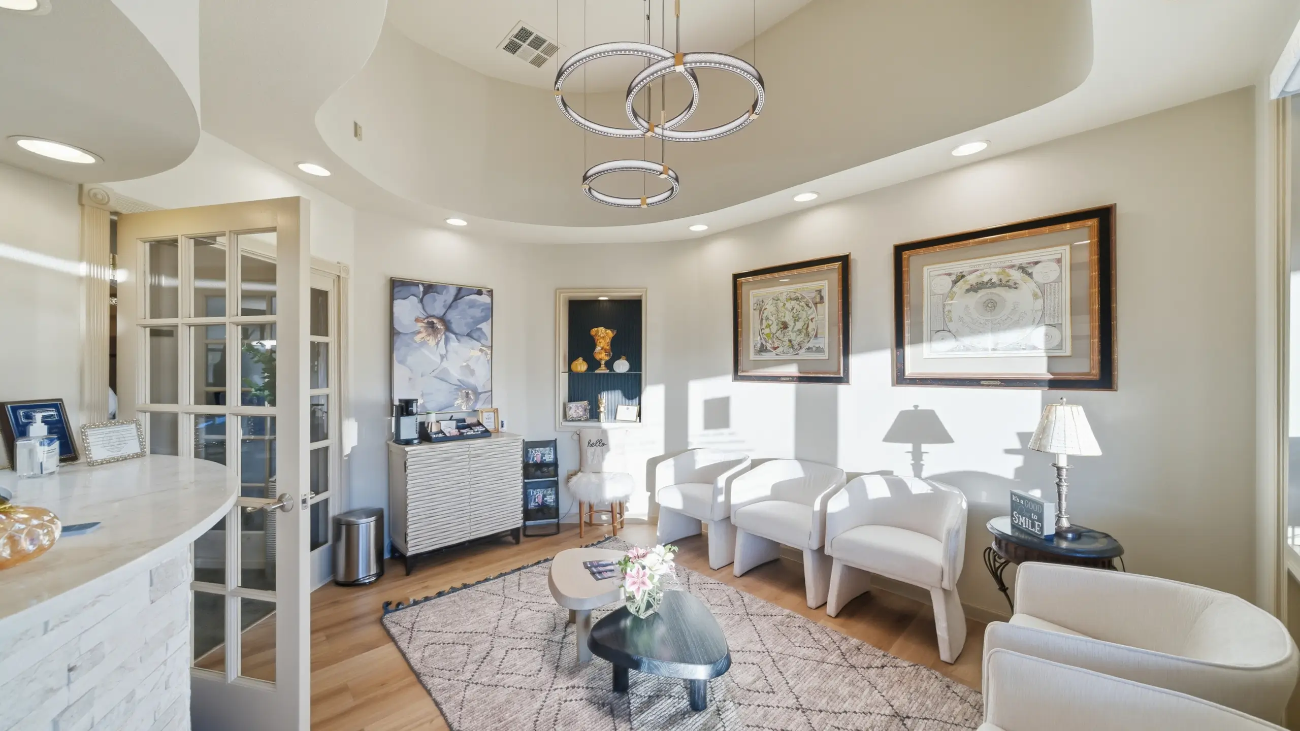 Bright, modern waiting room with white armchairs, a patterned rug, circular pendant lights, framed artwork, and a coffee station.