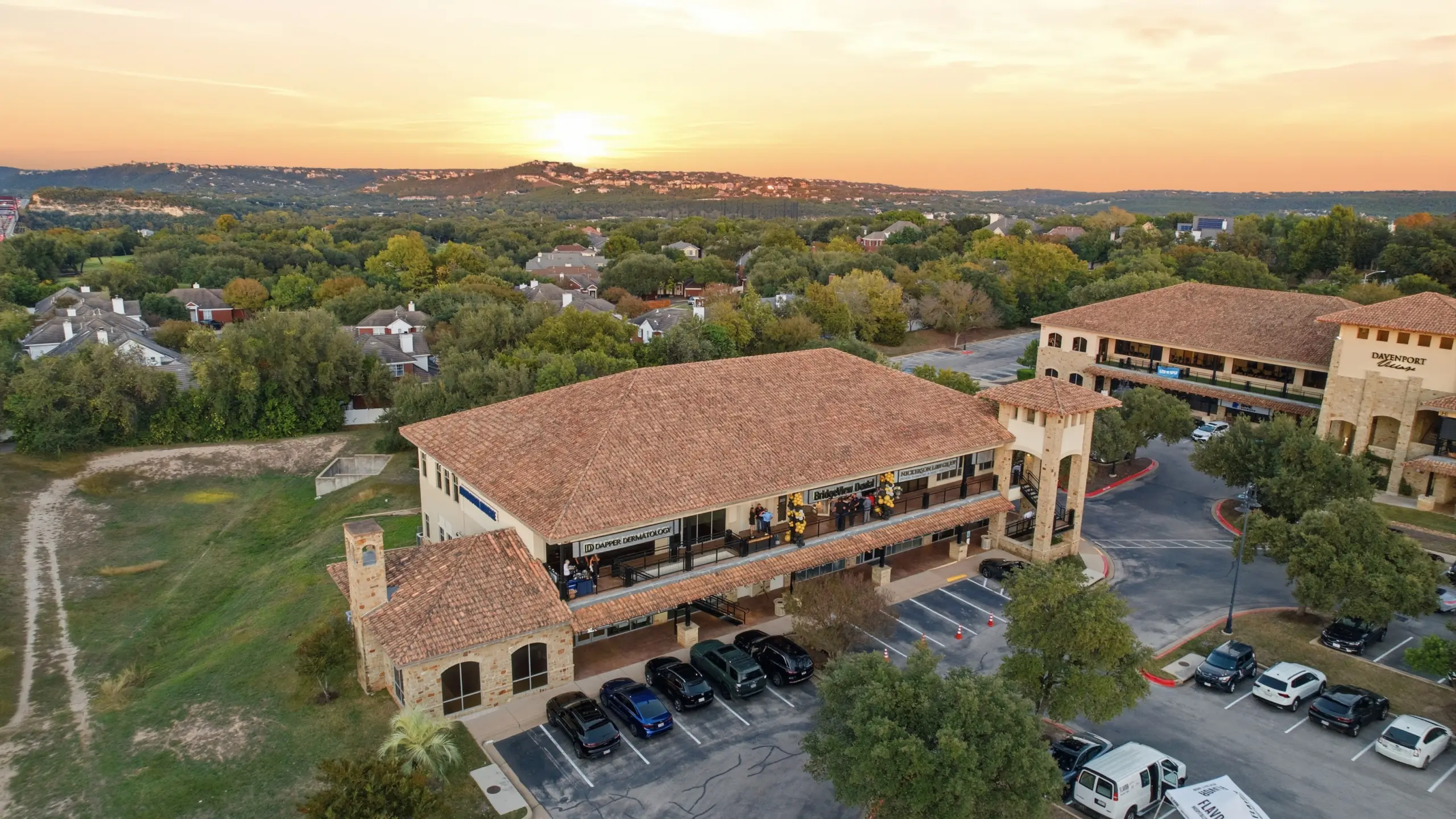 Aerial view of a commercial building complex with tiled roofs surrounded by trees and parked cars during a sunset.