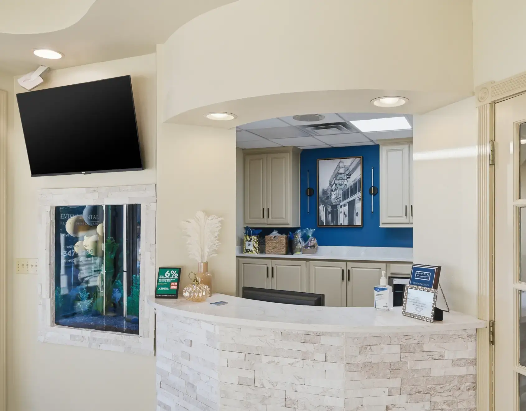 Reception desk area with white stone facade, decorative vase with feathers, hand sanitizer, and a wall-mounted TV above a small aquarium.