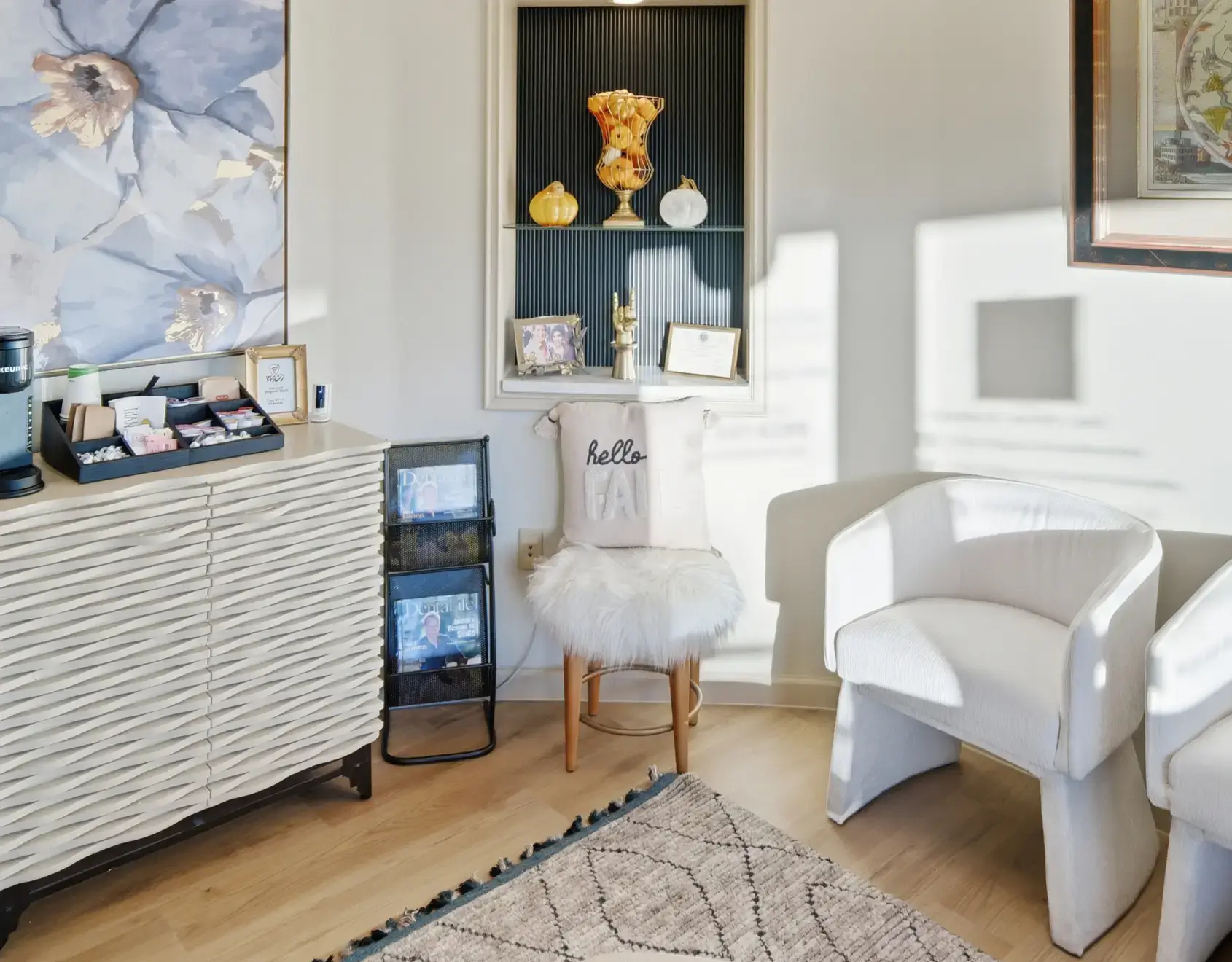 Sunlit cozy corner of a room featuring a white cushioned chair, a wooden stool with a fluffy seat and pillow, a textured cabinet with coffee supplies, a magazine rack, and decorative items including a floral wall painting and gold pumpkins on a built-in shelf.
