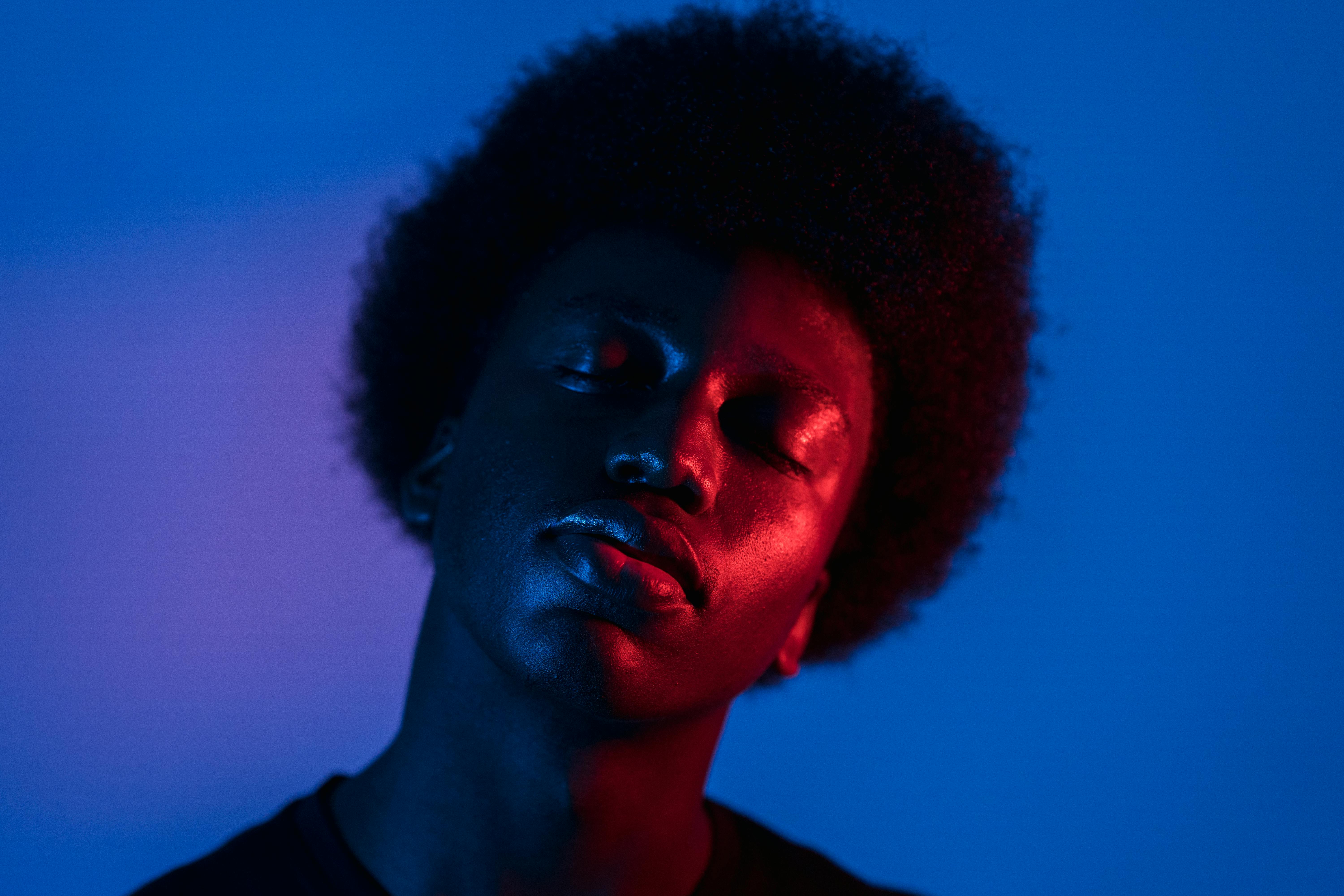 Portrait of a person with closed eyes illuminated by contrasting red and blue lighting against a blue background.