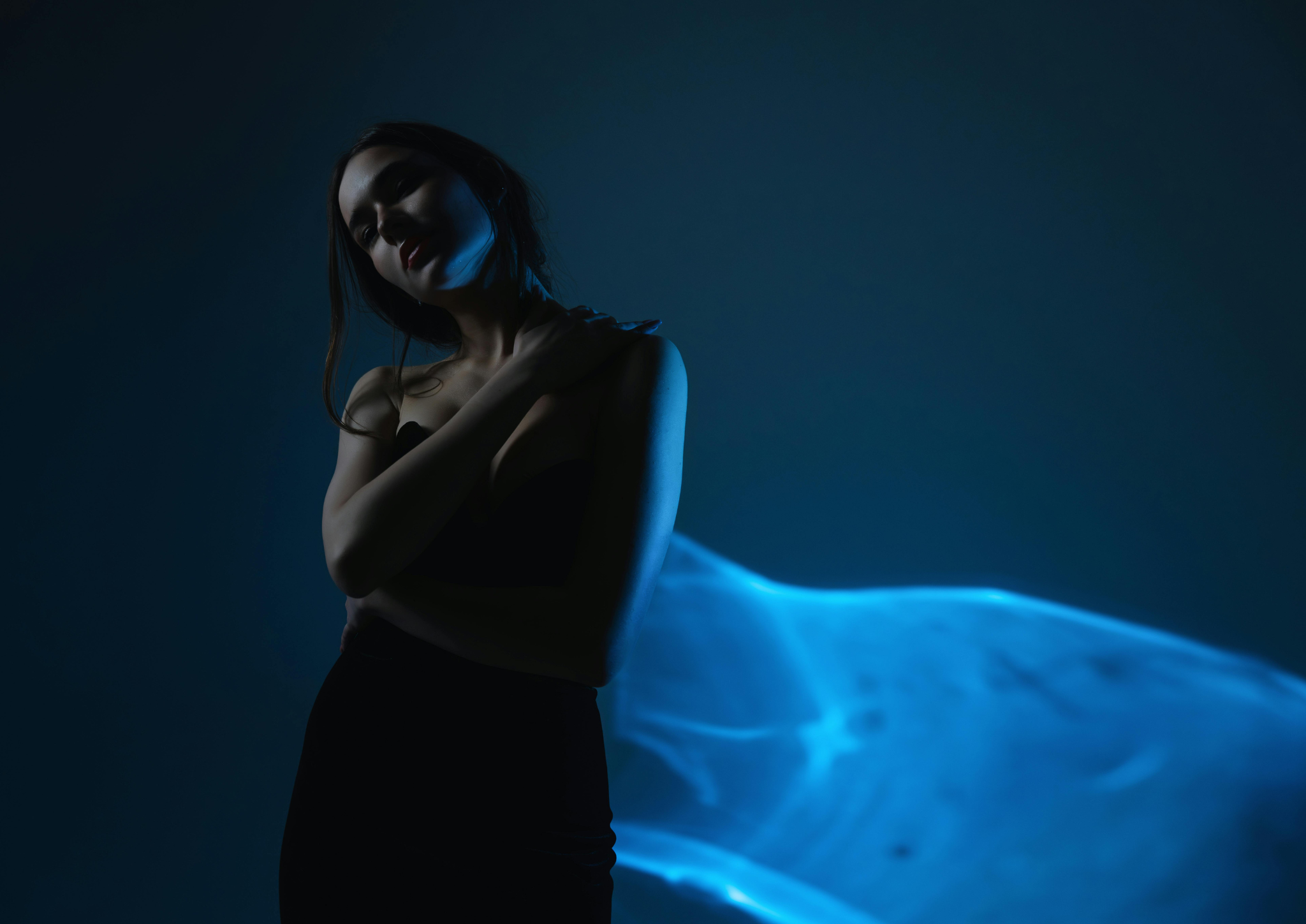 Woman in black dress with arms crossed against a dark background illuminated by soft blue light.