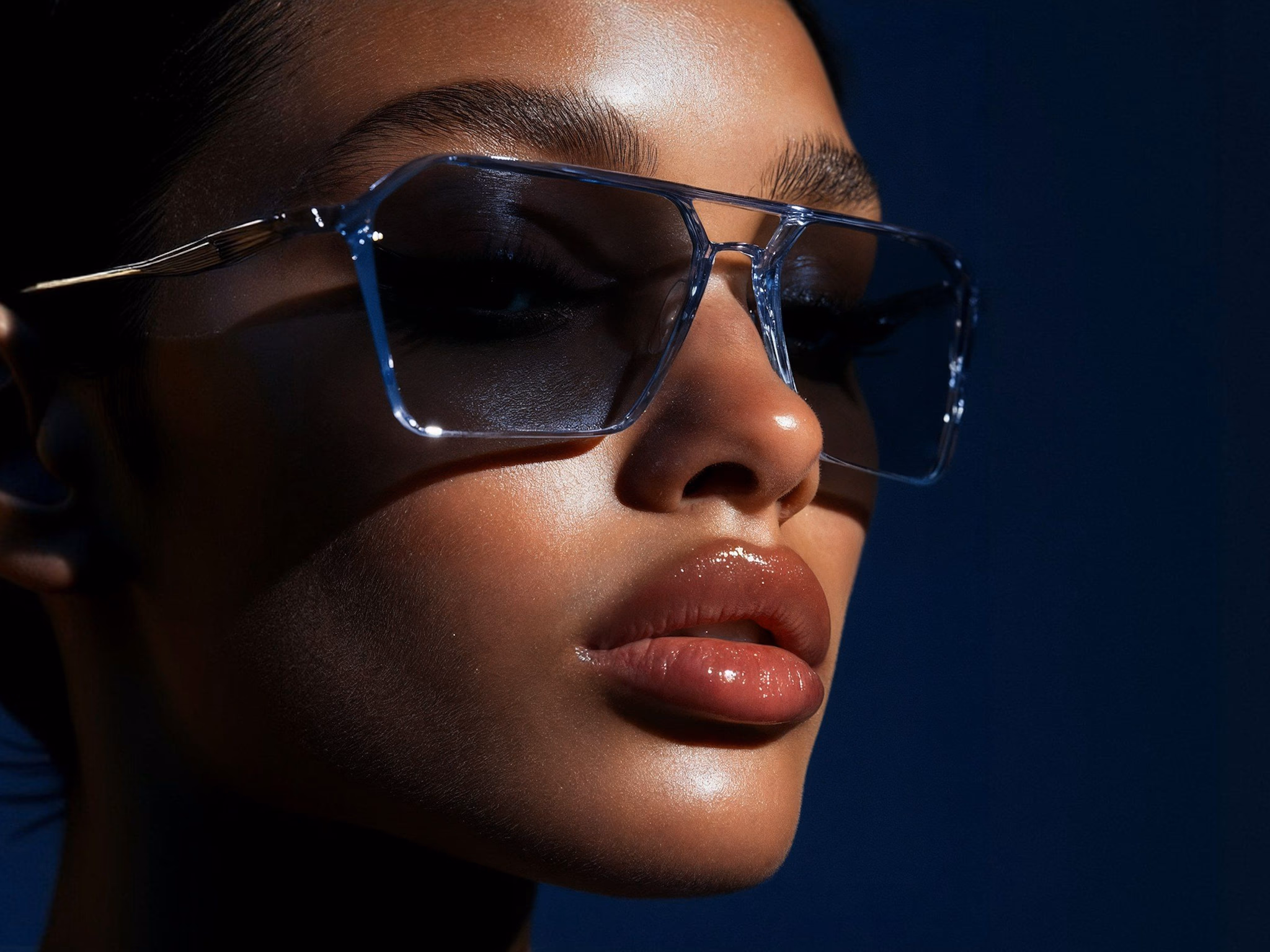 Close-up of a woman with glossy lips wearing large transparent sunglasses against a dark background.