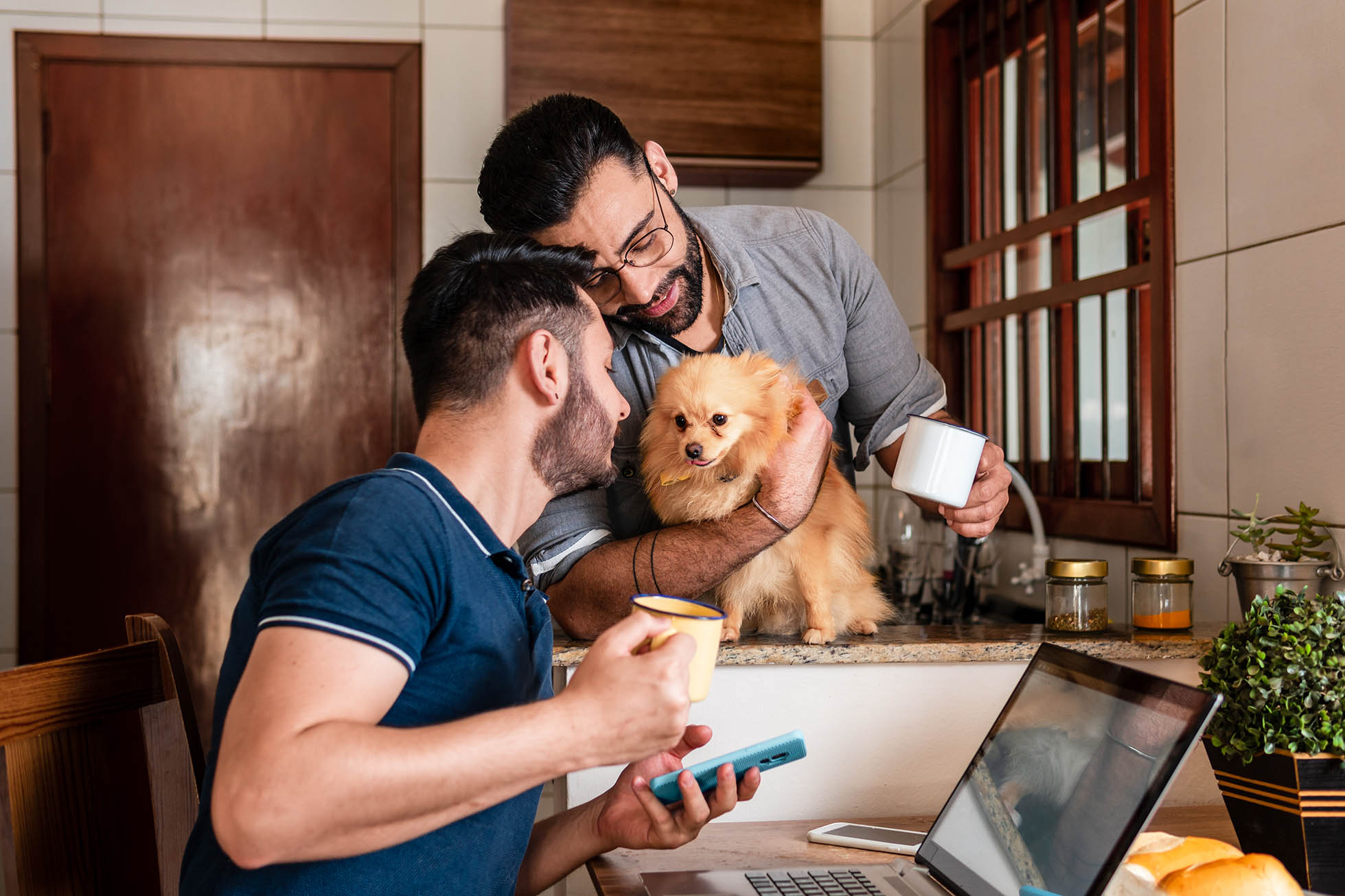 A young couple drinks coffee in their home. One is working on his computer while the other holds their dog.