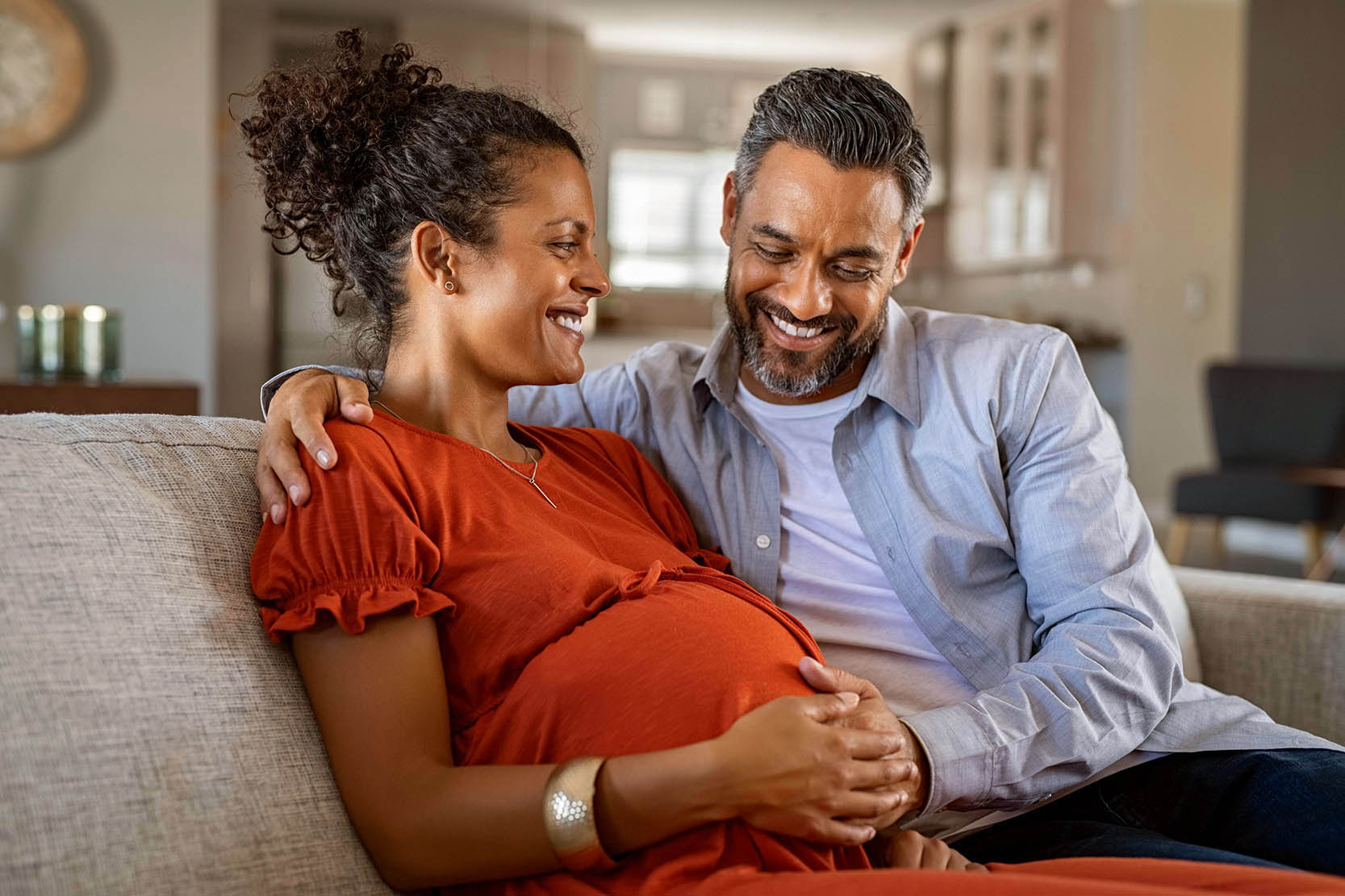 A young couple sits on their couch. The woman is pregnant, and they both have their hands on her belly and are smiling. 