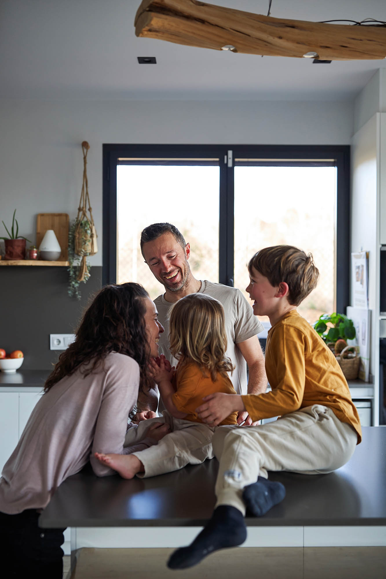 A family with two young kids laughing and talking together in the kitchen.  The kids are sitting on the table while the parents are standing and holding them. 