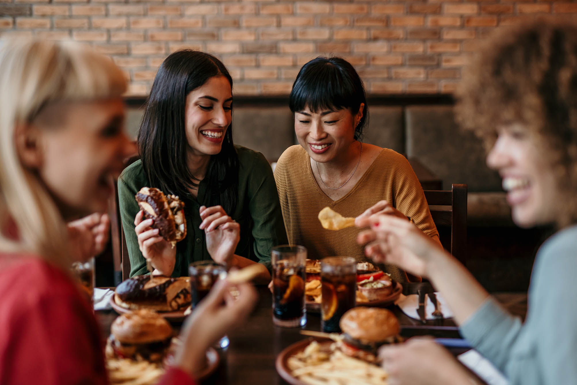 A diverse group of women enjoyed a meal together at a restaurant. Everyone is laughing and talking. There is a food and drinks on the table.
