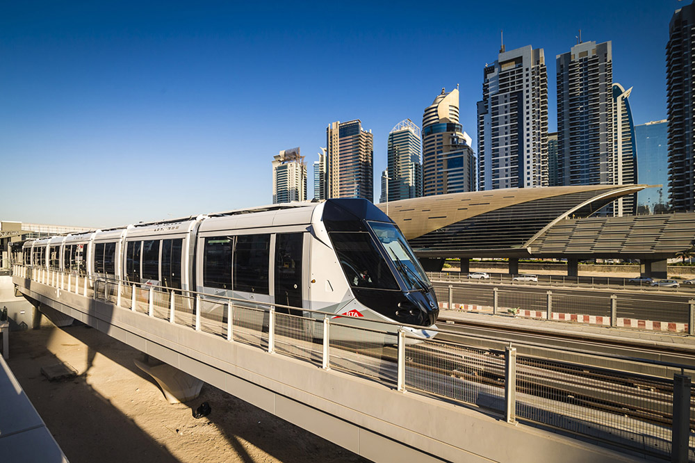 Modern white metro train on elevated tracks with a city skyline of tall buildings in the background under a clear blue sky.