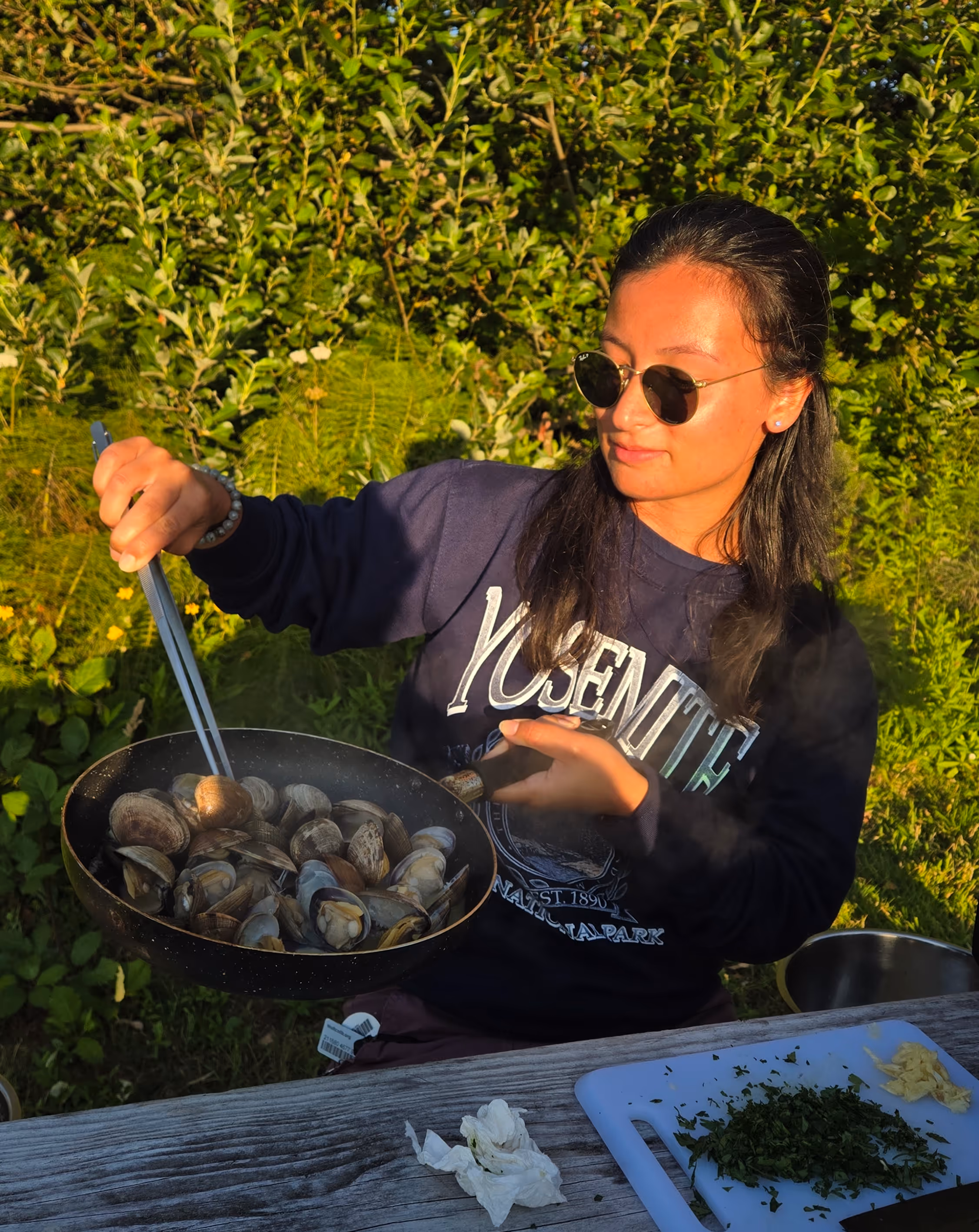 Person wearing sunglasses and a Yosemite sweatshirt cooking clams in a pan outdoors with green foliage in the background.