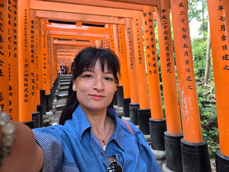 Woman taking a selfie in front of a pathway lined with bright orange Torii gates with Japanese inscriptions.