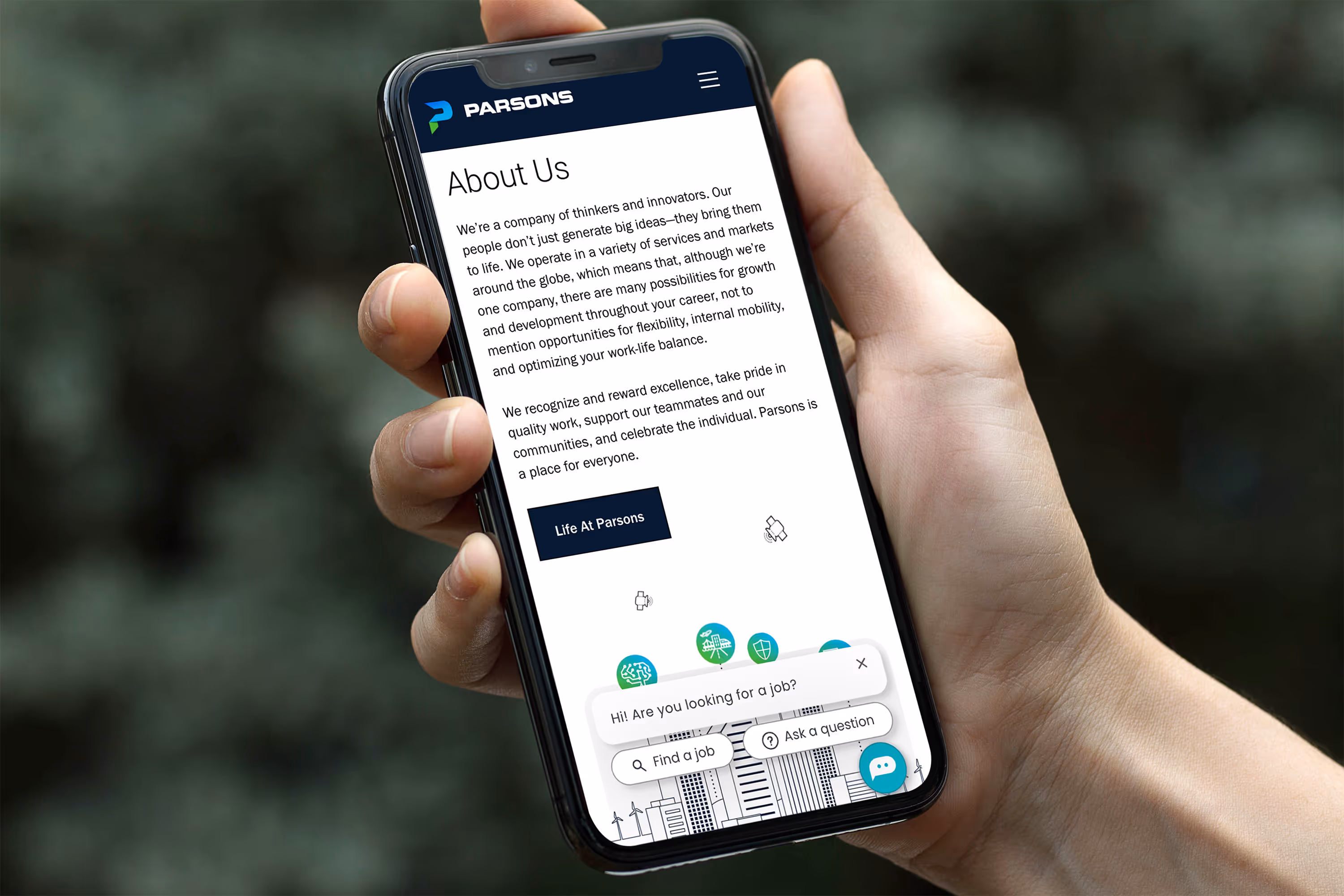 Hand holding a smartphone displaying Parsons company About Us page with text about company values and a chat popup offering job search help.