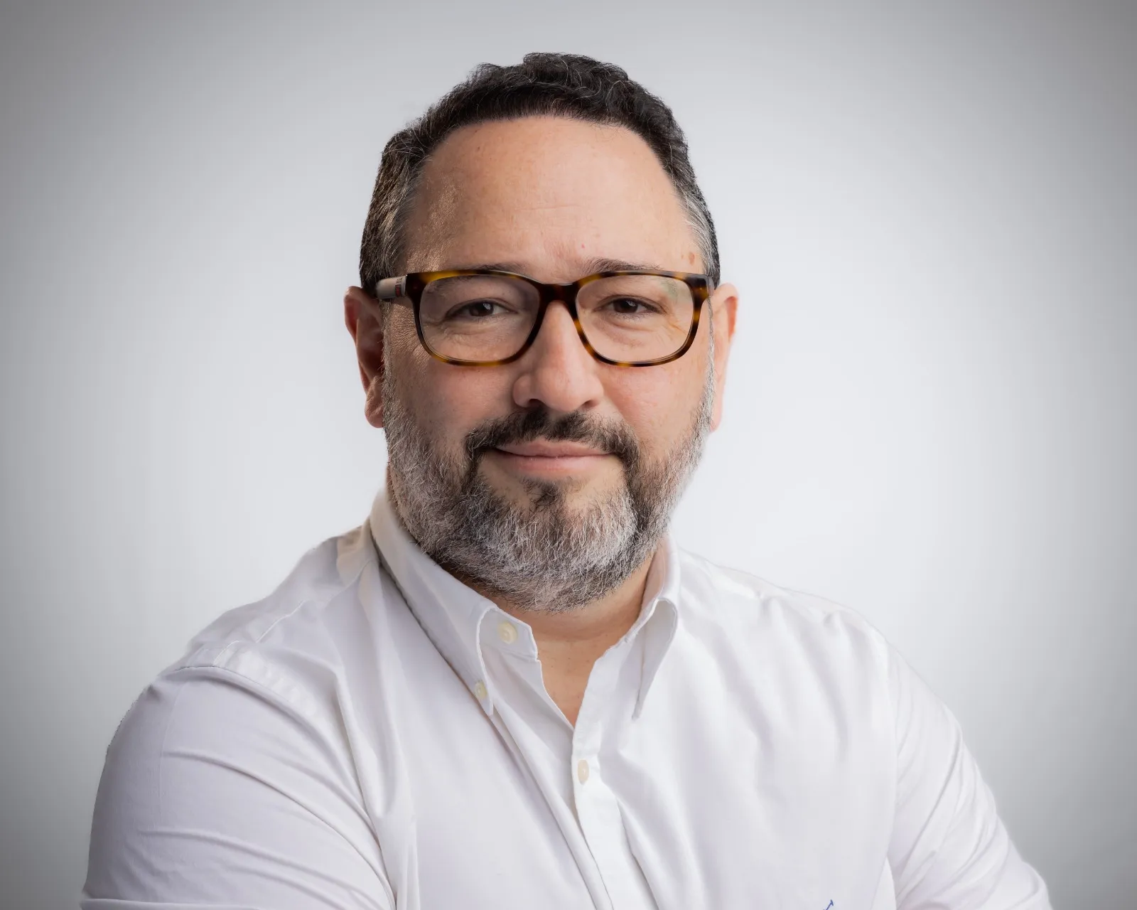 Middle-aged man with dark hair, beard, glasses, and white shirt smiling against a plain gray background.