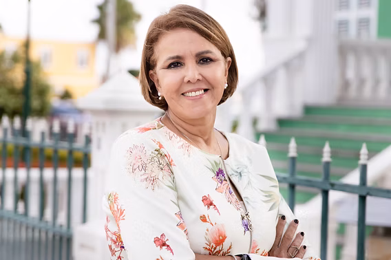 Smiling woman in floral jacket with arms crossed standing outdoors by a fence and white building.