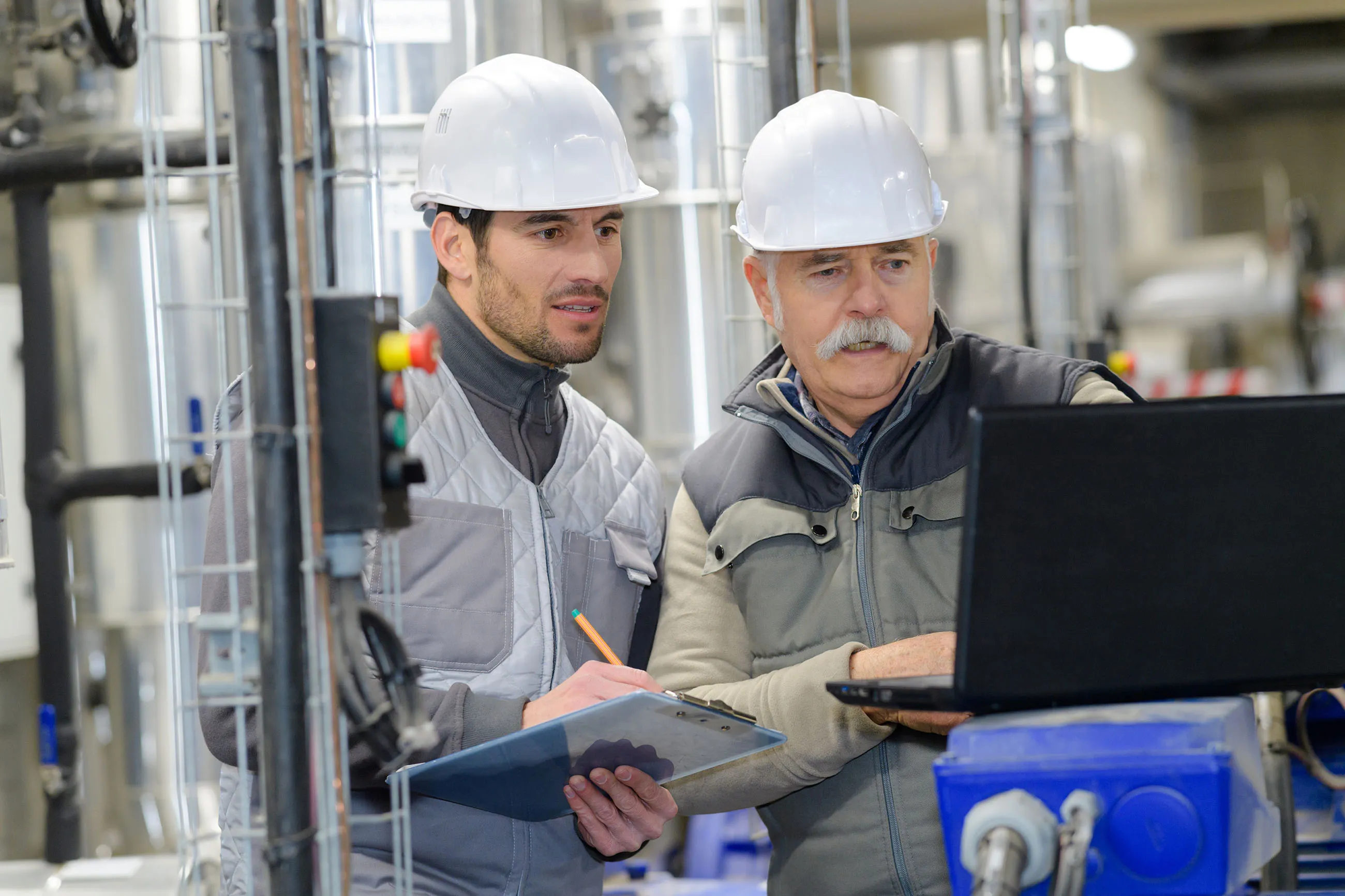Two male engineers wearing white hard hats and work jackets examining a laptop and clipboard in an industrial setting.