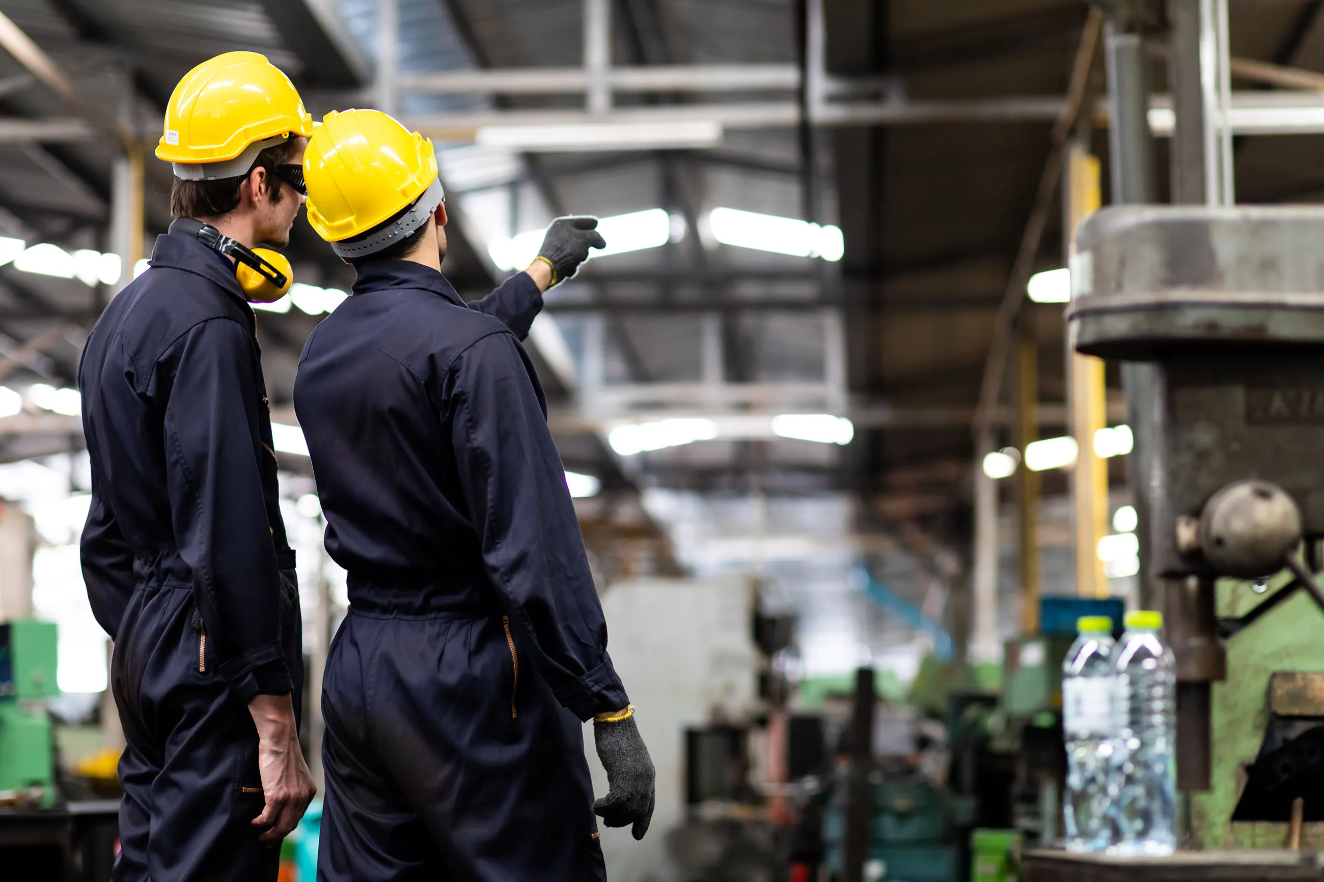 Two factory workers wearing yellow safety helmets and dark uniforms discuss inside an industrial workshop.