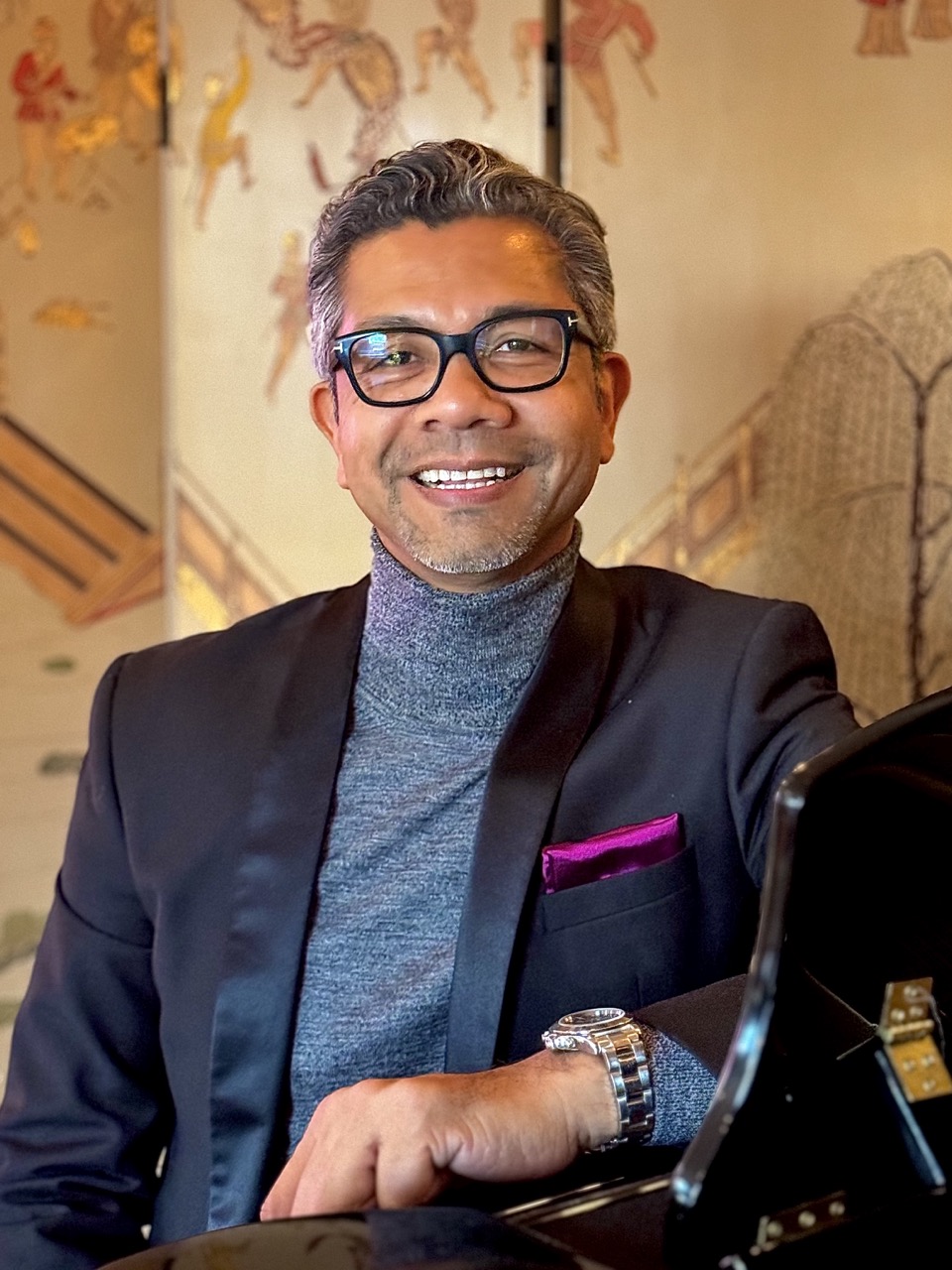 Portrait of interior designer Will Brown wearing a black blazer and gray turtleneck, smiling while seated indoors.