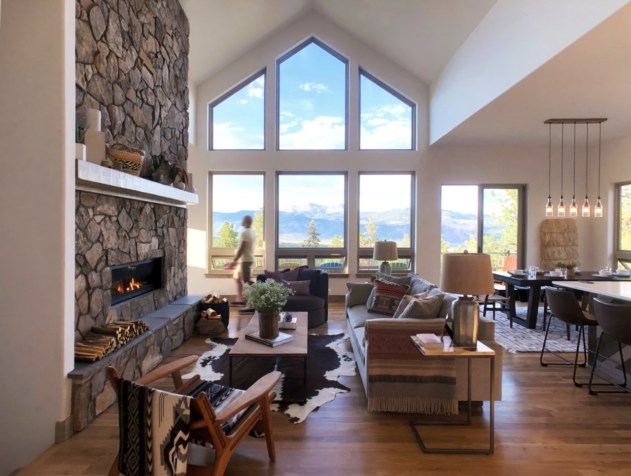Mountain great room with tall windows, stone fireplace, wood floors, and panoramic mountain views.