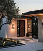 Modern house entrance with wooden door, illuminated wall sconces, and landscaped steps at dusk.