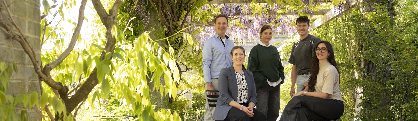 Das Team von der Geschäftsstelle posiert lächelnd in einem grünen Garten mit blühender Pergola.