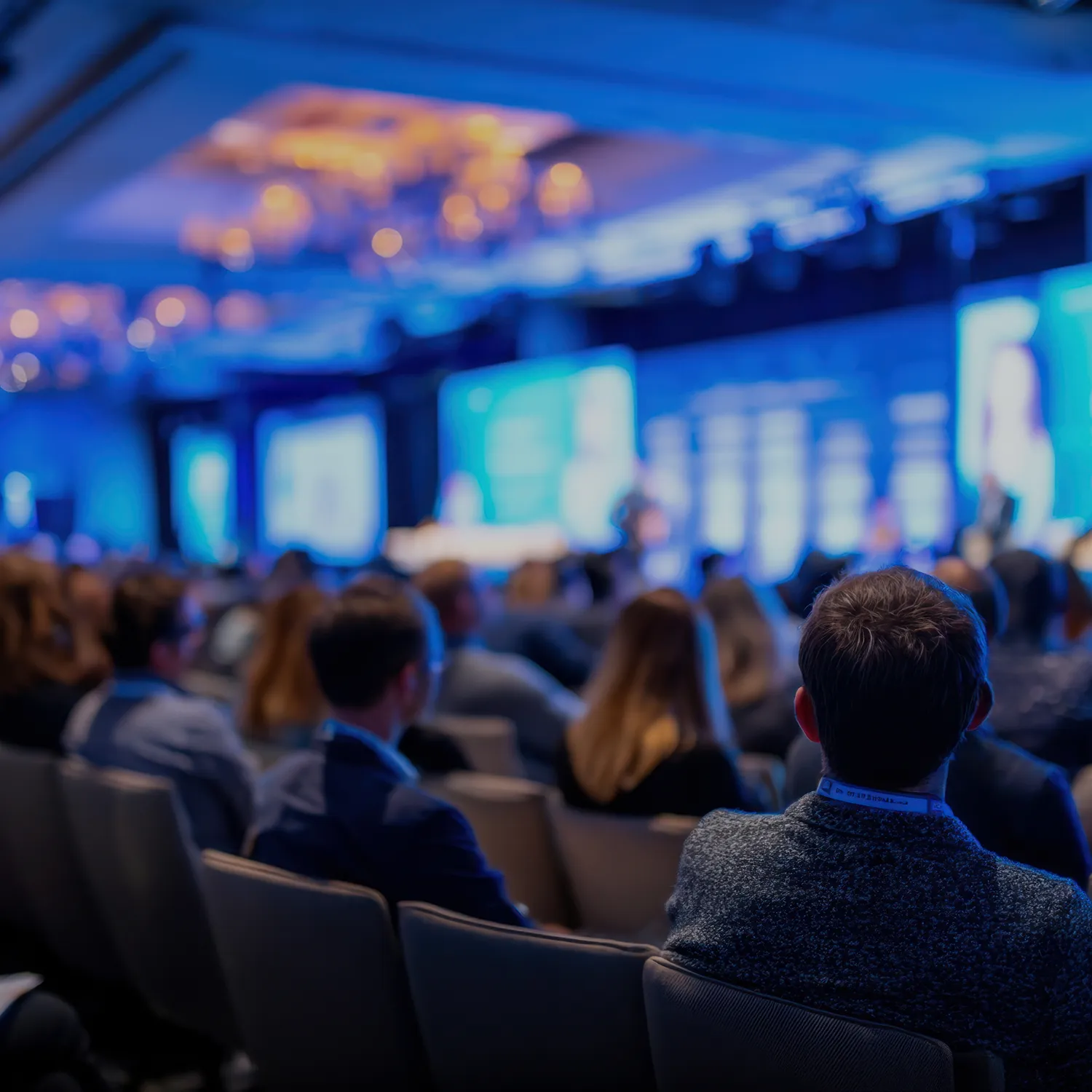 Audience seated facing a stage with large blue-lit screens at a conference or seminar.