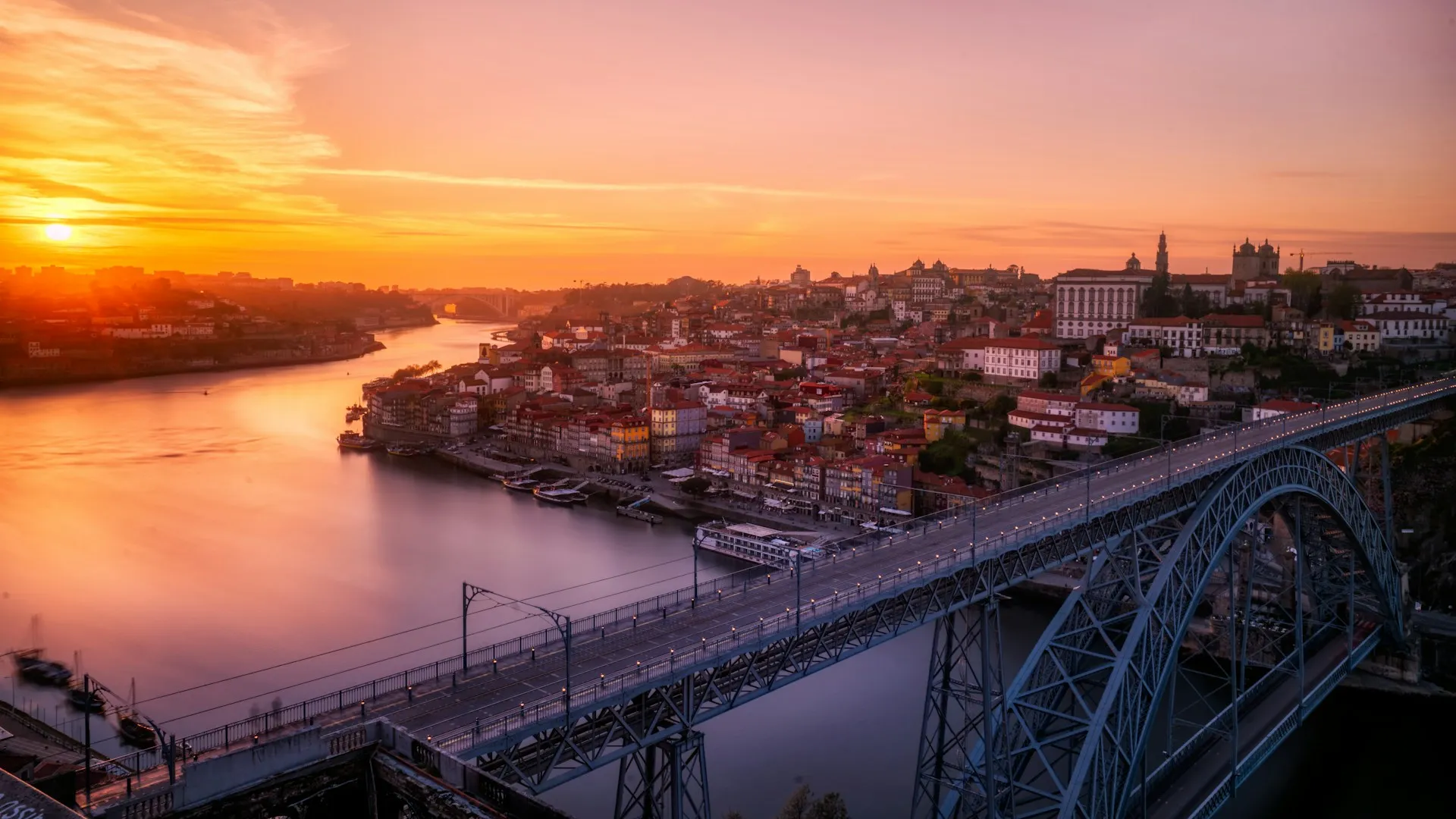 Sunset over river with arched metal bridge and city buildings glowing in warm light.