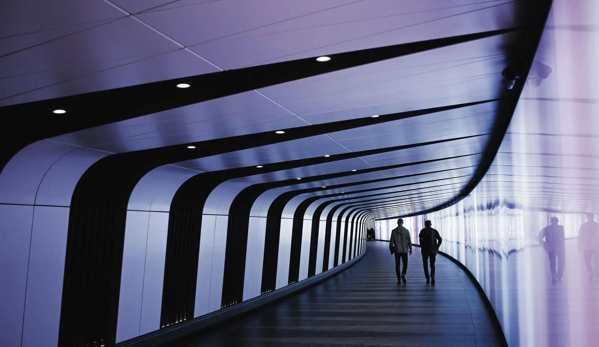 Two people walking in a modern curved corridor with black and white walls and ceiling lights.