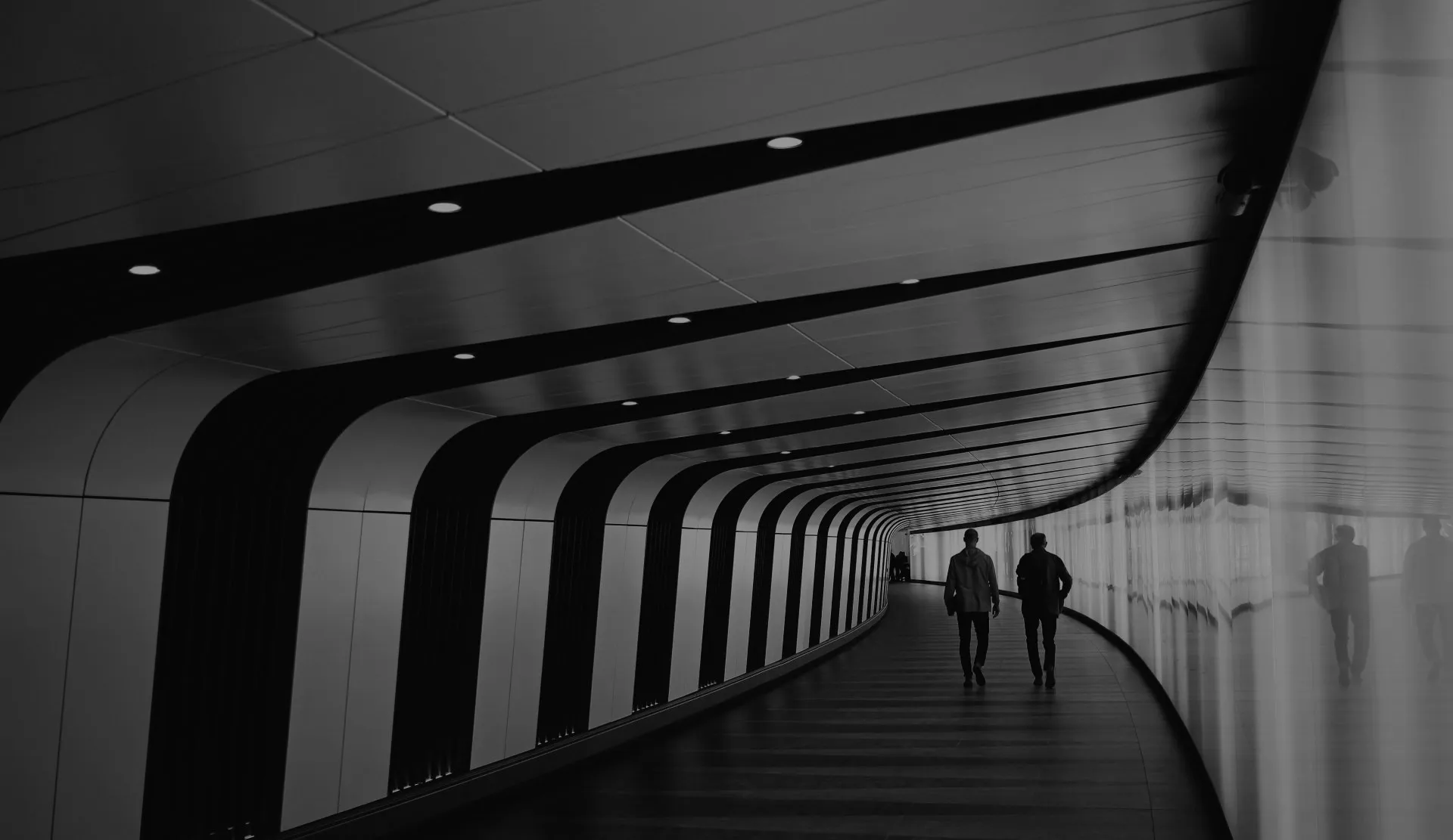 Two people walking through a curved tunnel with black beams and reflective wall on the right.