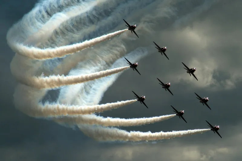 Nine aircraft flying in formation creating curved smoke trails against a cloudy sky.
