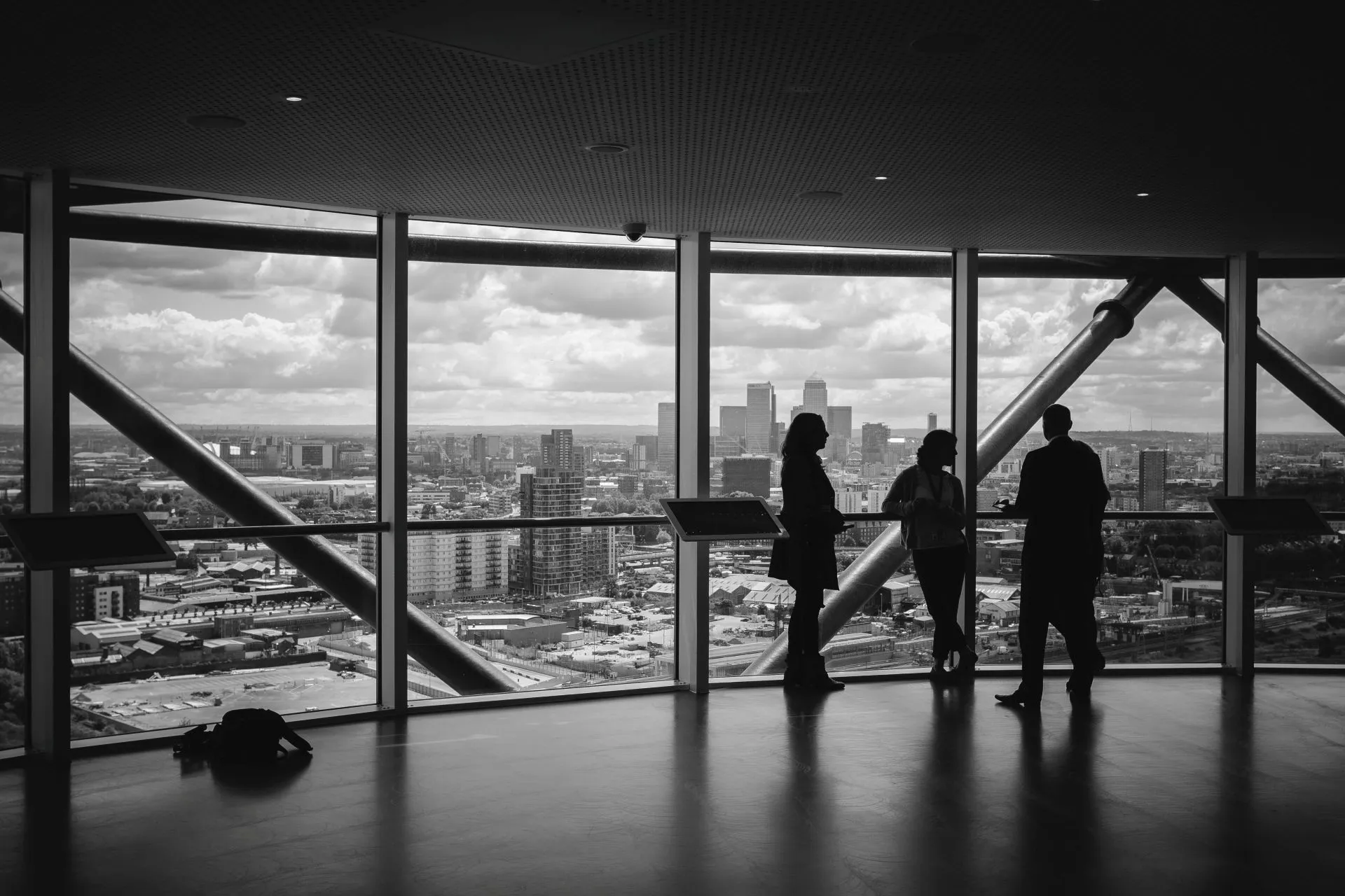 Silhouettes of three people standing near large windows overlooking a cityscape with cloudy sky.
