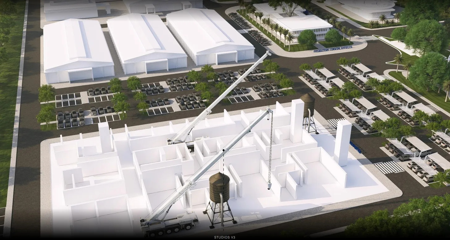 Aerial view of a white building under construction with cranes, parking lots, trees, and other buildings.