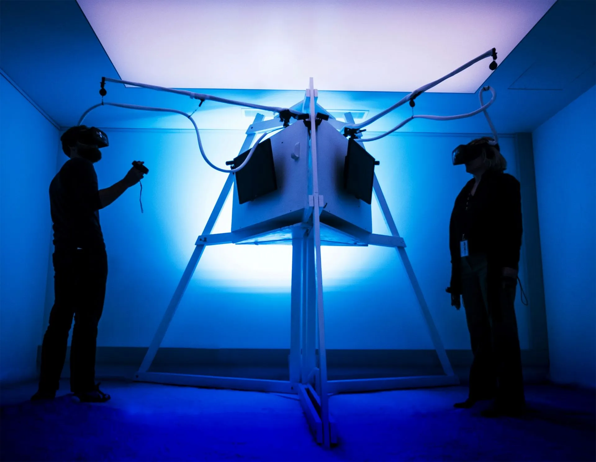 Two people wearing VR headsets interacting with a cube-shaped VR setup in a blue-lit room.