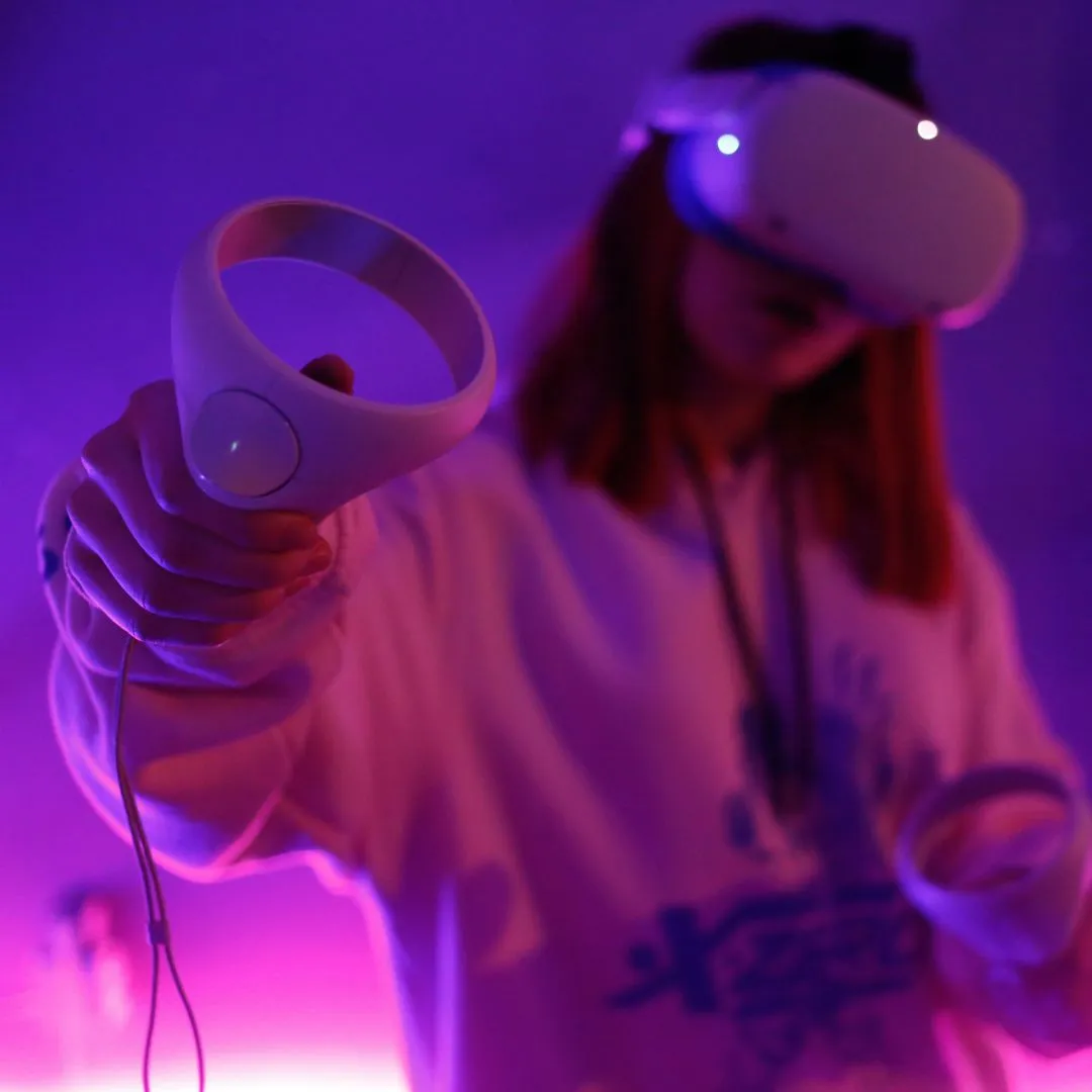 Person wearing a VR headset and holding VR controllers in purple-lit room.