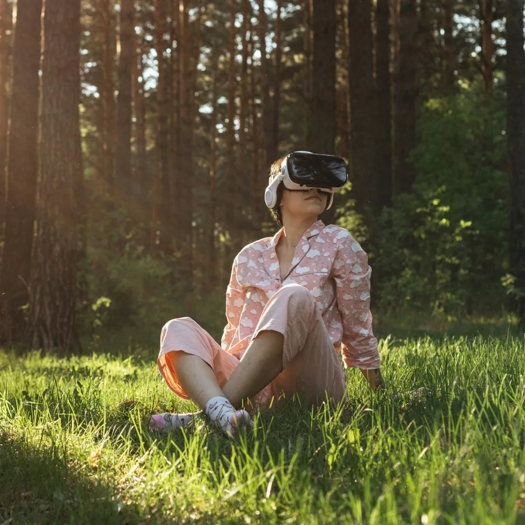 Person sitting on grass in forest wearing pink pajamas and a virtual reality headset with headphones.