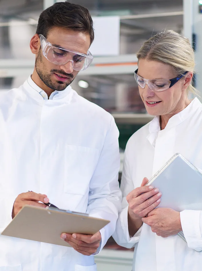Two scientists in lab coats and safety glasses reviewing a clipboard and tablet