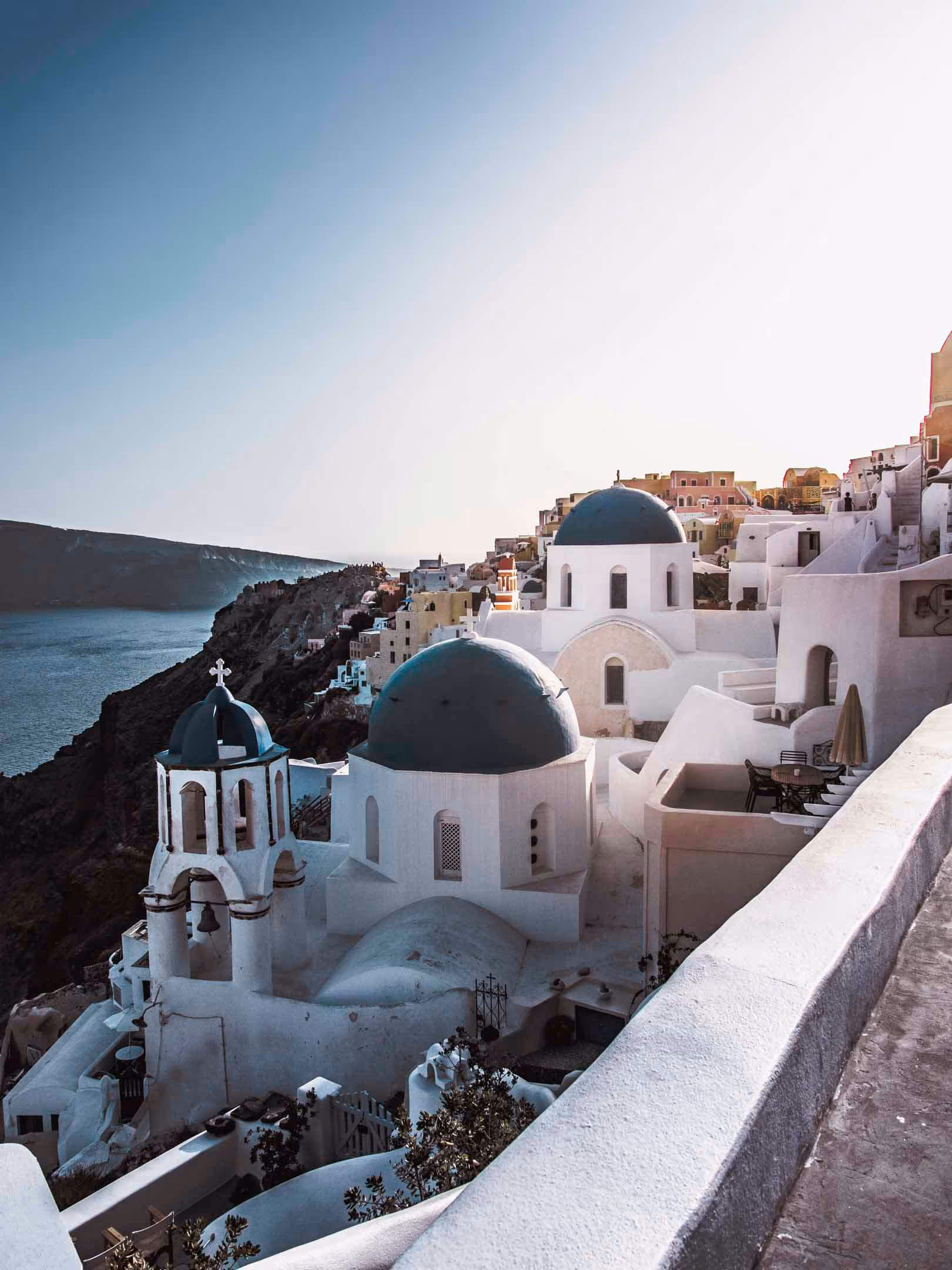 White buildings with blue domes and a bell tower on a cliffside overlooking the sea at sunset in Santorini, Greece.