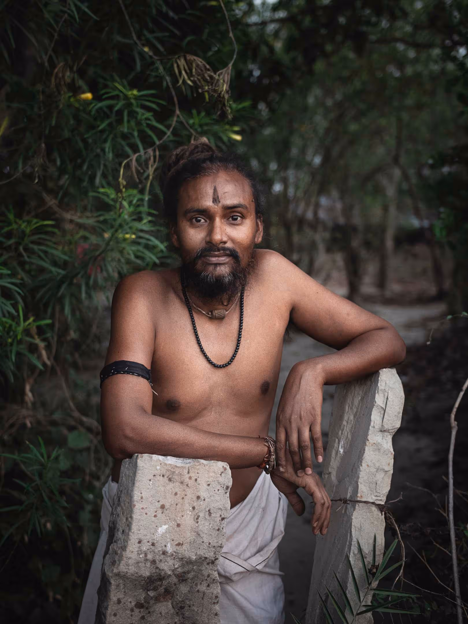 A shirtless man with a beard and a necklace leans on two stone pillars in a forest setting.