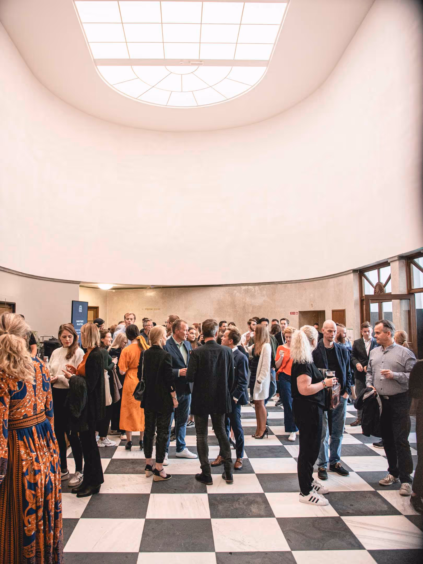 Group of people socializing in a room with a large skylight and black-and-white checkered floor.
