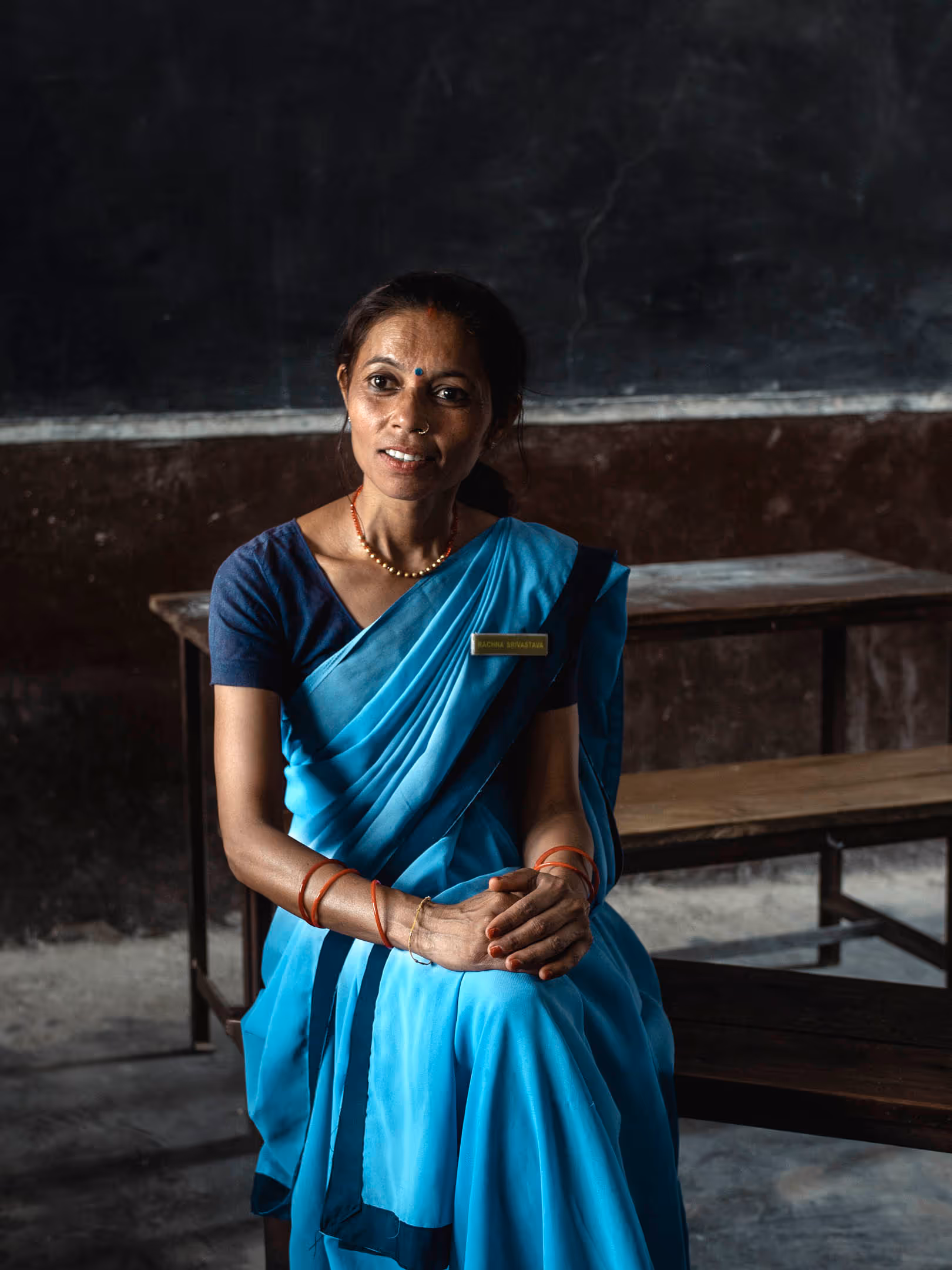 Smiling woman in a blue sari sitting indoors with folded hands and a classroom desk behind her.