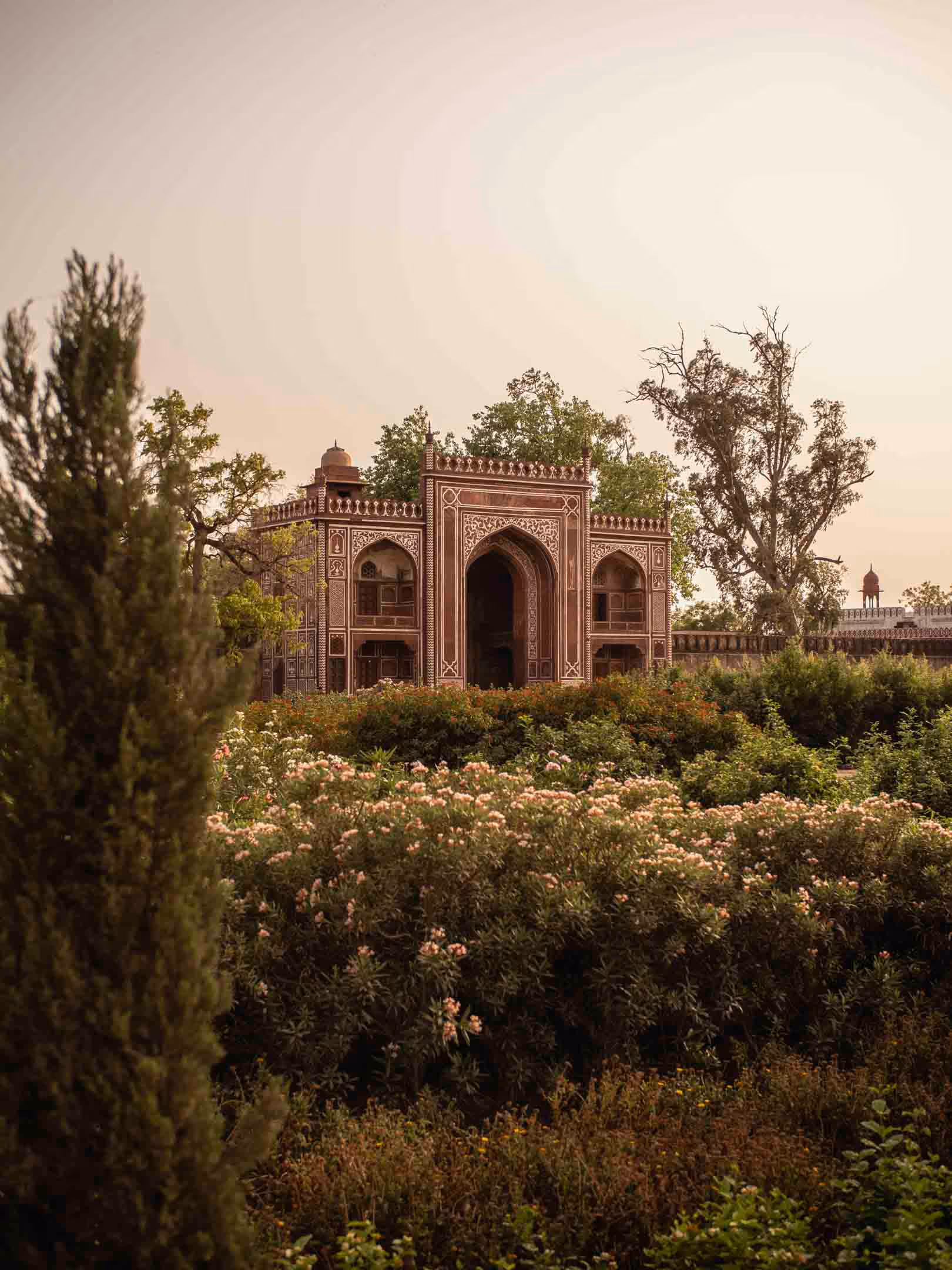 Ornate historic building with intricate patterns behind lush garden bushes and tall trees under a soft golden sky.
