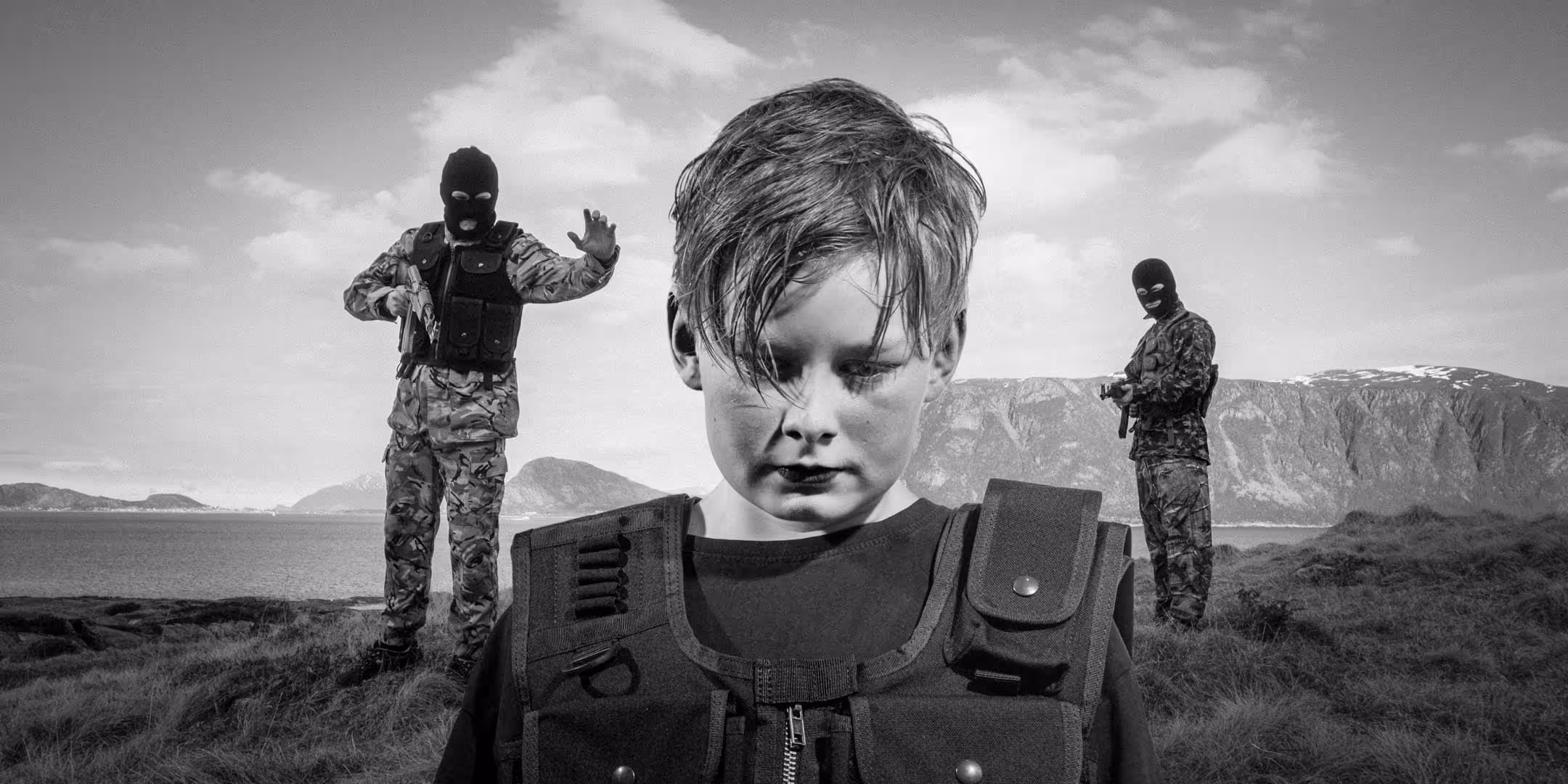 Close-up of a young boy wearing a tactical vest looking down with two armed masked soldiers in camouflage standing behind him outdoors near water and mountains.