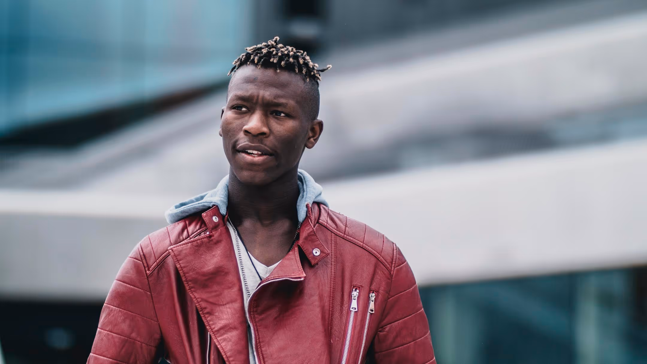 Young man with short twisted hair wearing a red leather jacket with a gray hooded sweatshirt underneath, standing outdoors in an urban setting.