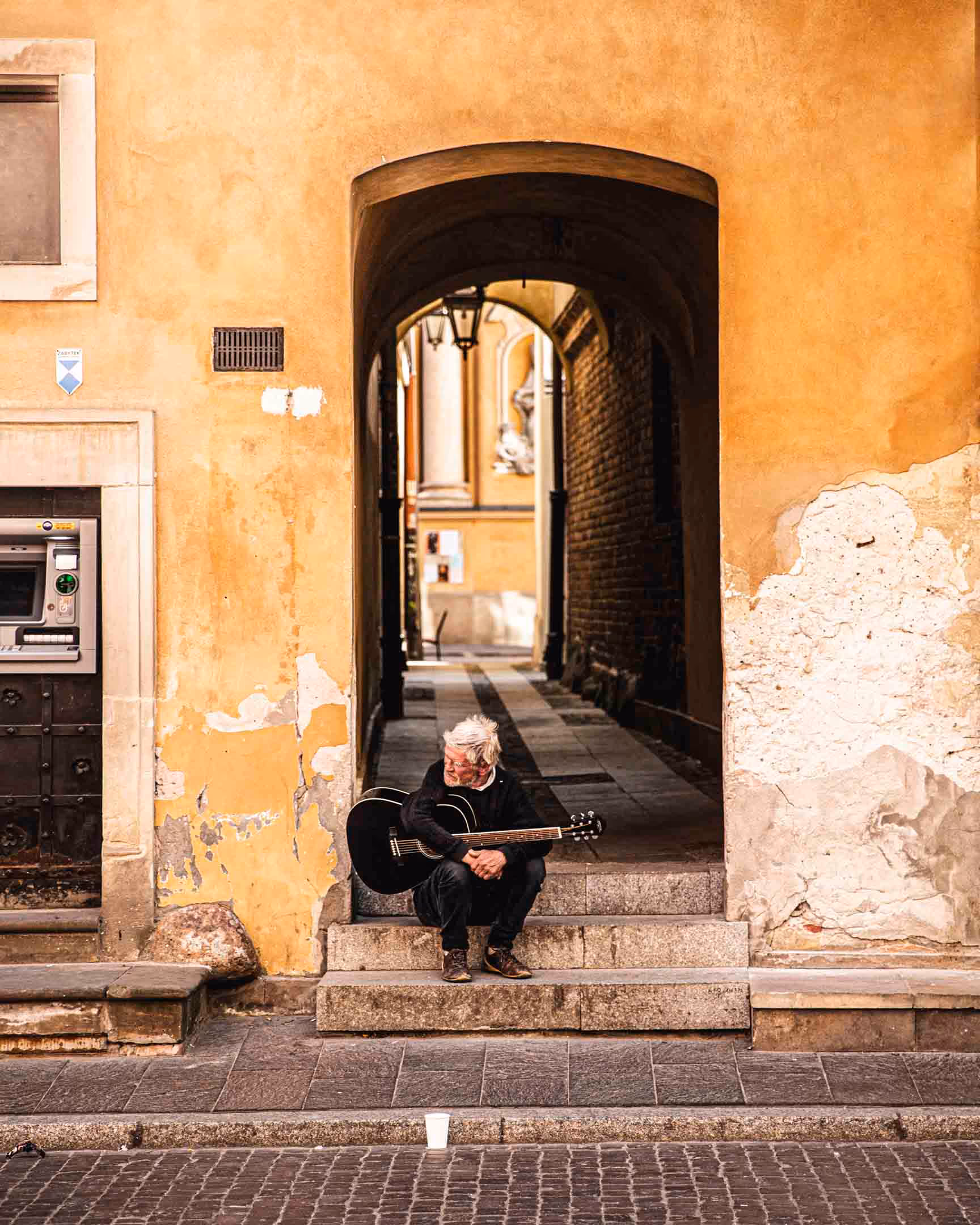 Elderly man with gray hair sitting on stone steps under an archway, holding a black acoustic guitar.