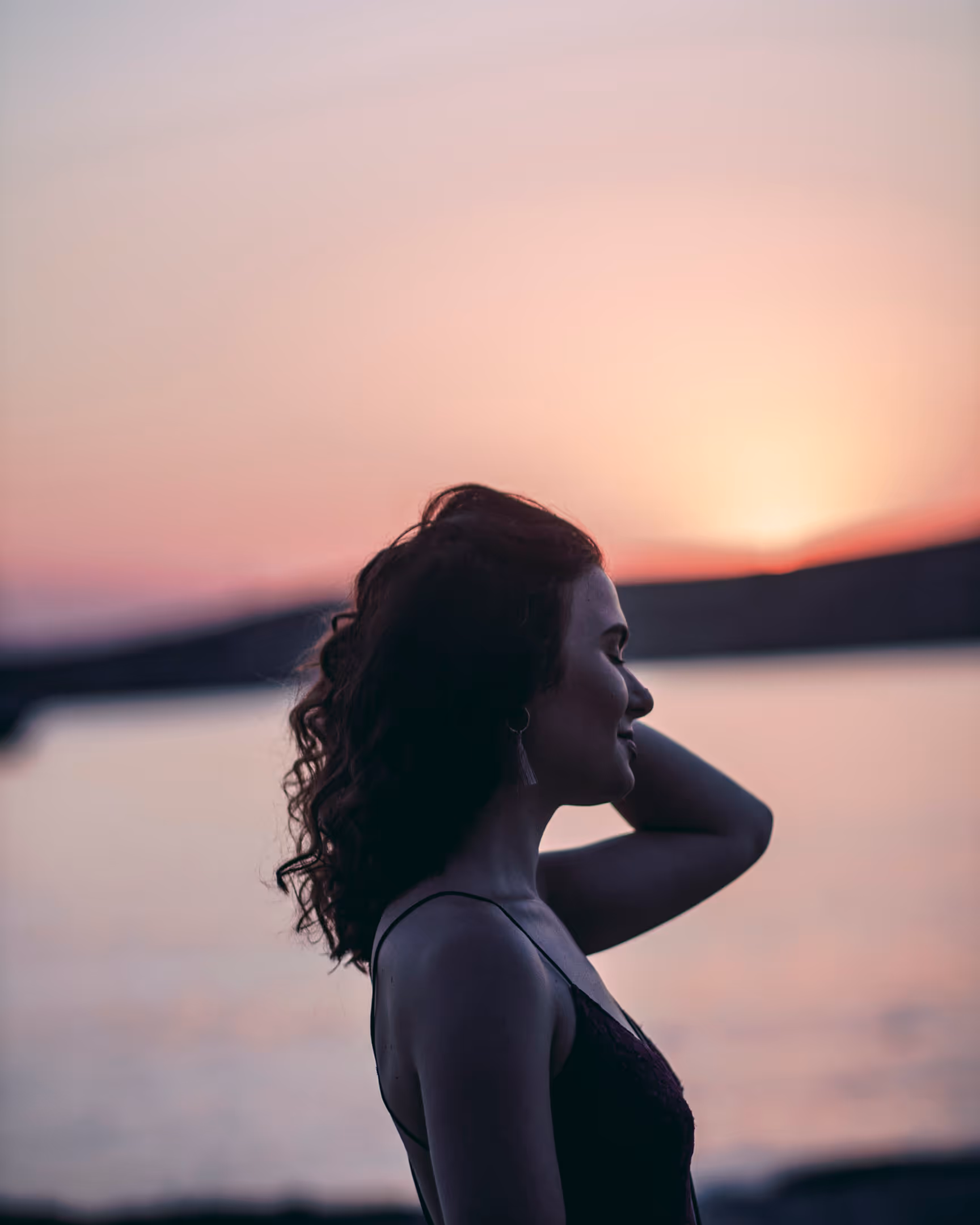 Silhouette of a woman with curly hair standing by water at sunset with eyes closed and hand behind head.