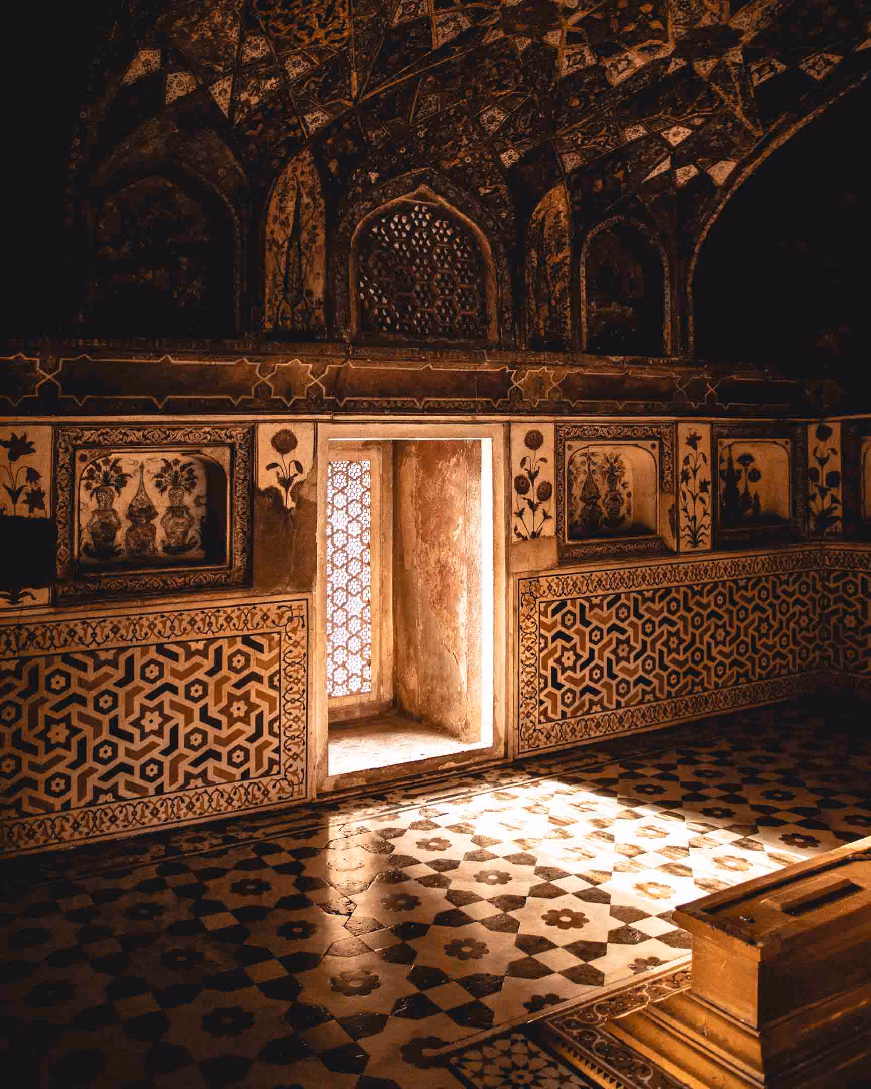 Dimly lit room with intricately patterned walls and floor, featuring a doorway illuminated by sunlight.