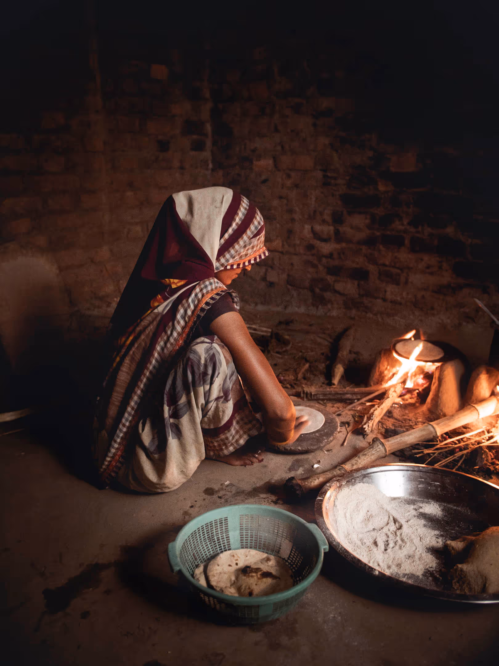 Woman in traditional attire making flatbread by a wood-fired stove inside a rustic kitchen.