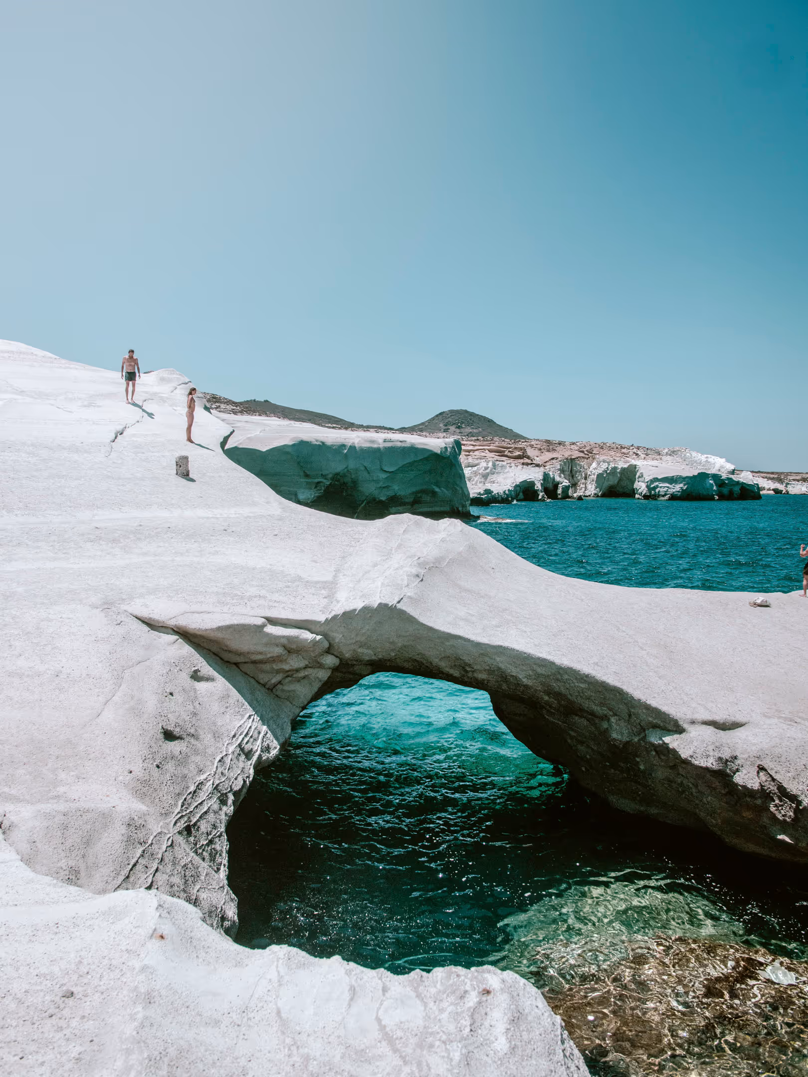 White rocky coastal arch over turquoise sea water with people standing on the rocks under a clear blue sky.
