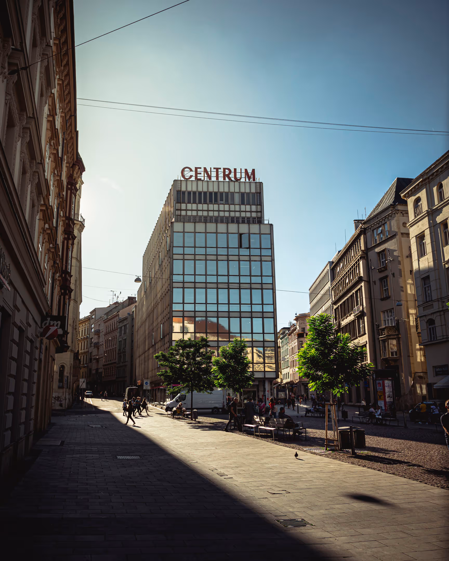 Busy city street with modern glass building labeled 'CENTRUM' and people walking and sitting on benches.